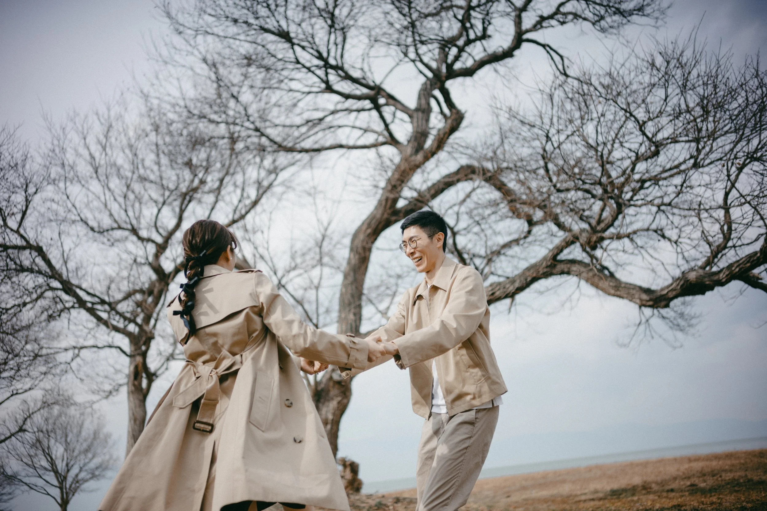 A young man and woman holding hands and smiling outdoors near a leafless tree on a cloudy day.