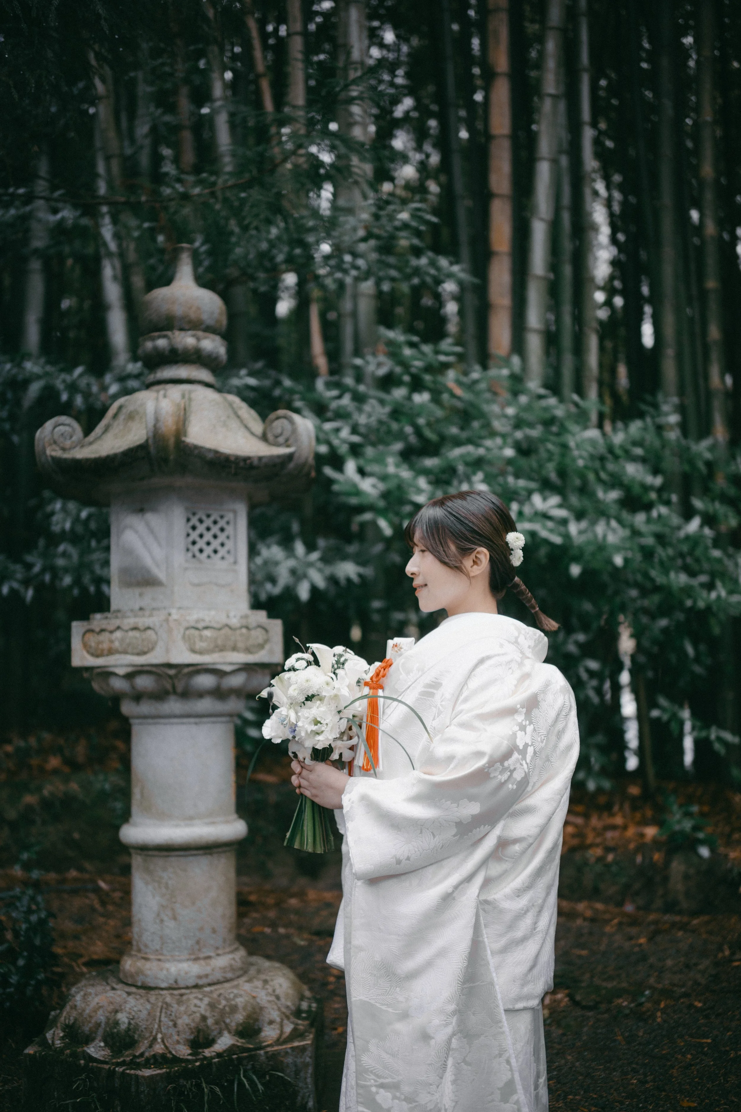 A woman in a traditional white wedding kimono holding a bouquet in a forested area near a stone lantern.