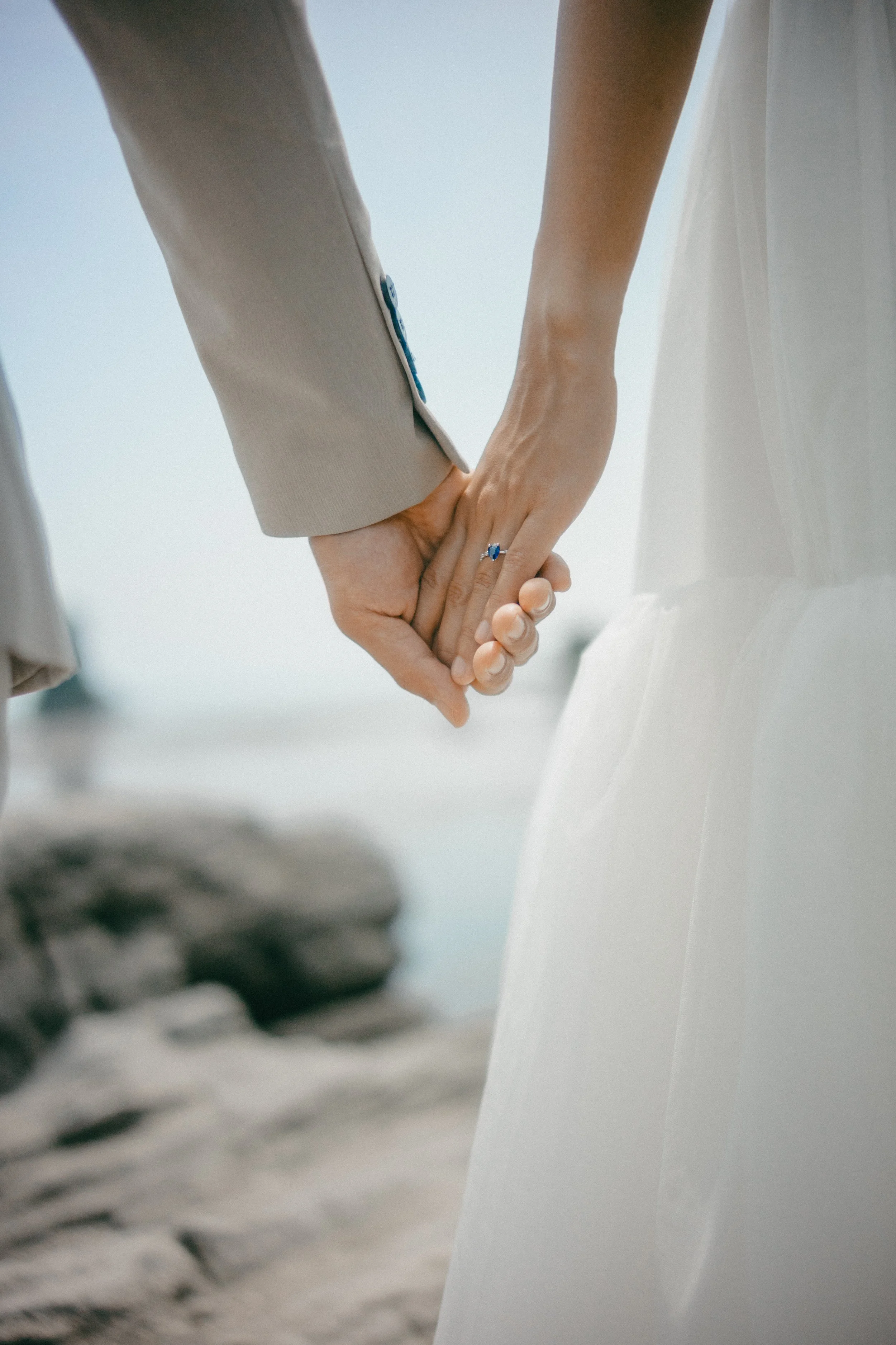 Close-up of a bride and groom holding hands, with the bride wearing a white dress and the groom in a beige suit, outdoors near rocks or water.