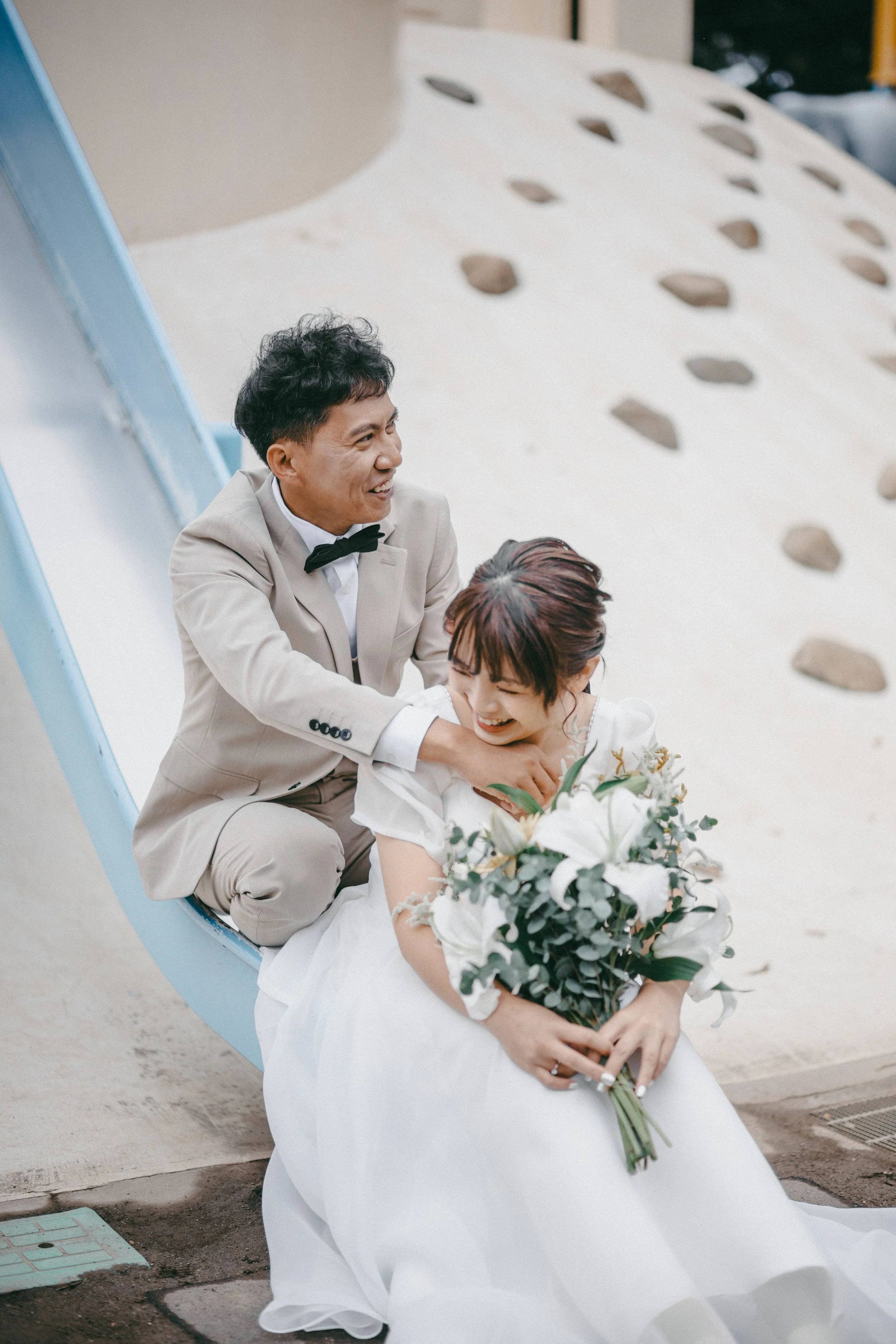 A bride and groom are playfully posing on a slide, with the groom in a beige suit and bow tie, and the bride in a white wedding dress holding a bouquet of white lilies and greenery. They are laughing and enjoying a fun moment