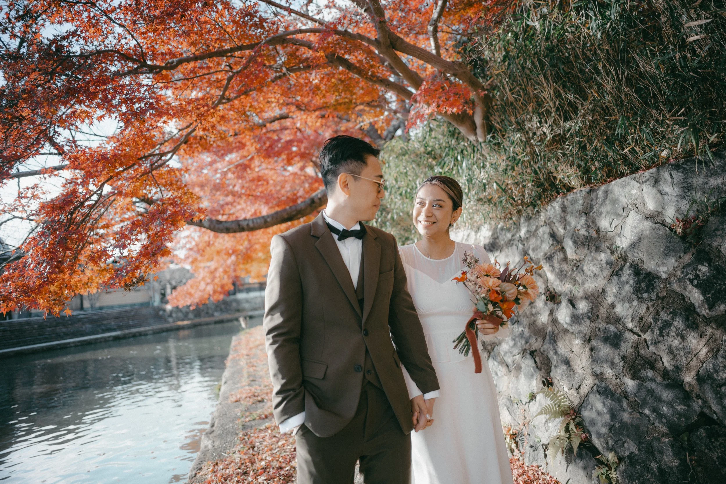 A couple dressed in wedding attire holding hands and smiling at each other outdoors during autumn, with orange-red leaves on trees and a body of water nearby.