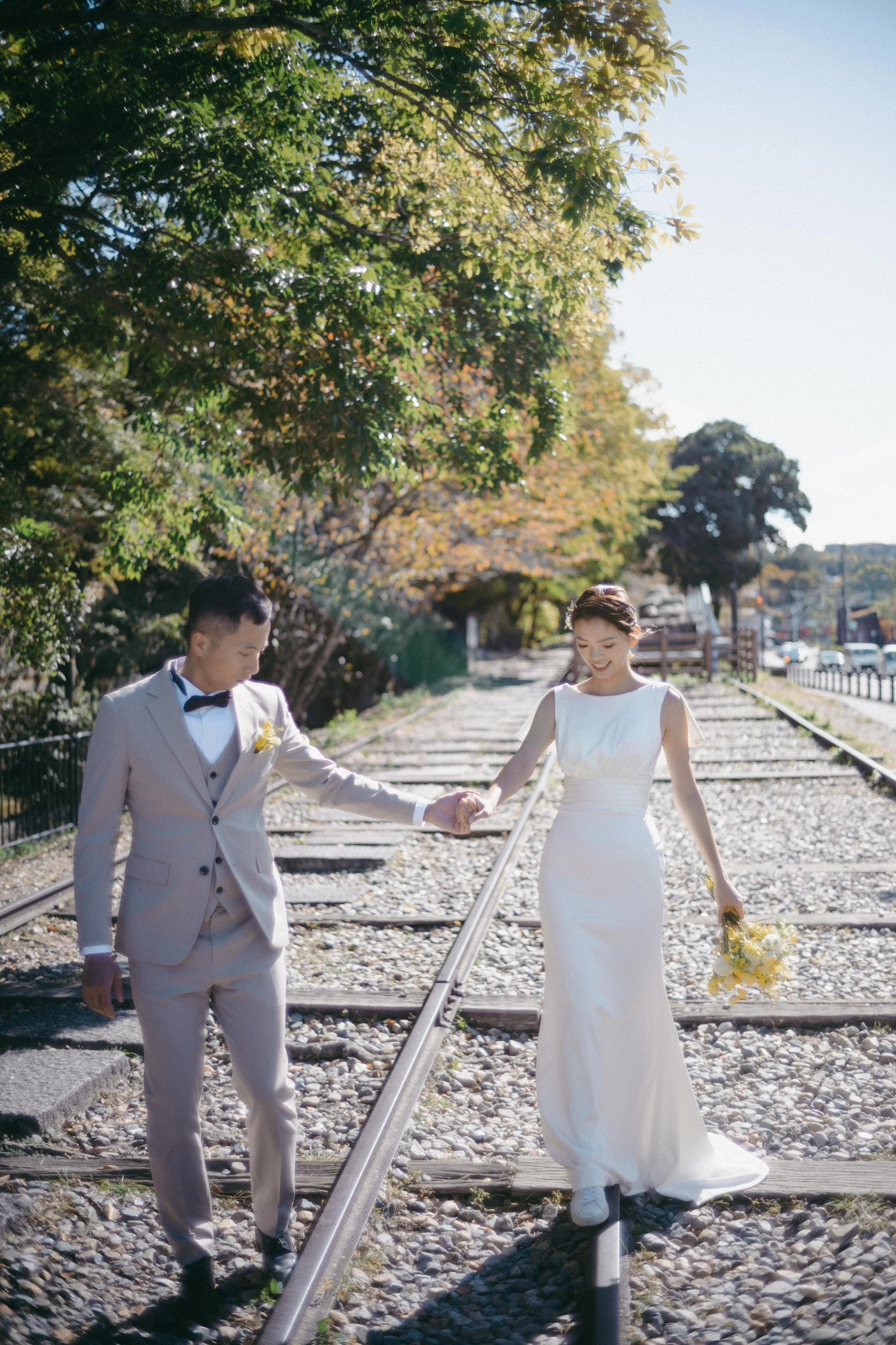 A bride and groom holding hands while walking on train tracks outdoors, with trees and a clear sky in the background.