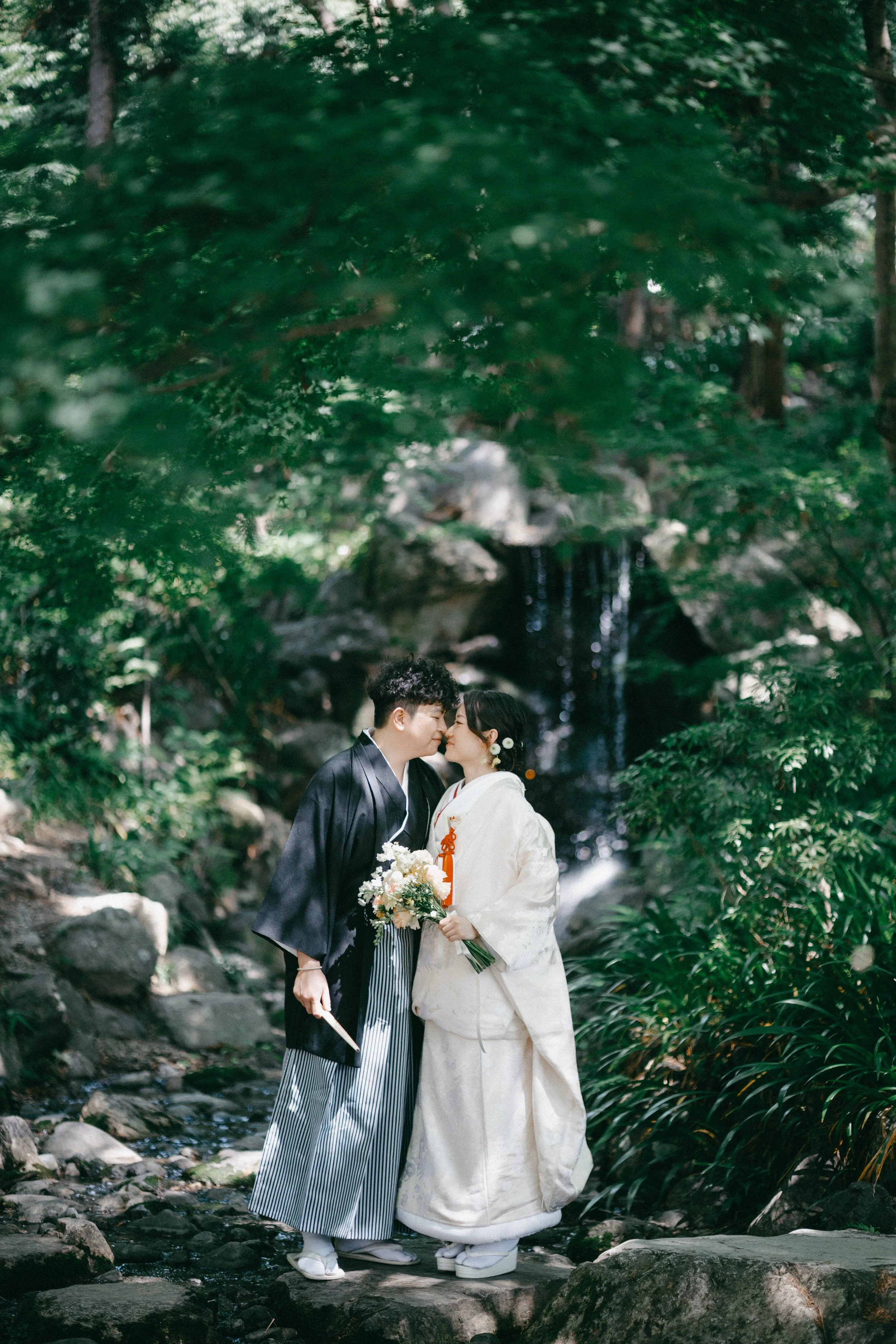 A couple in traditional Japanese wedding attire standing on rocks near a small waterfall in a lush green forest, about to kiss.