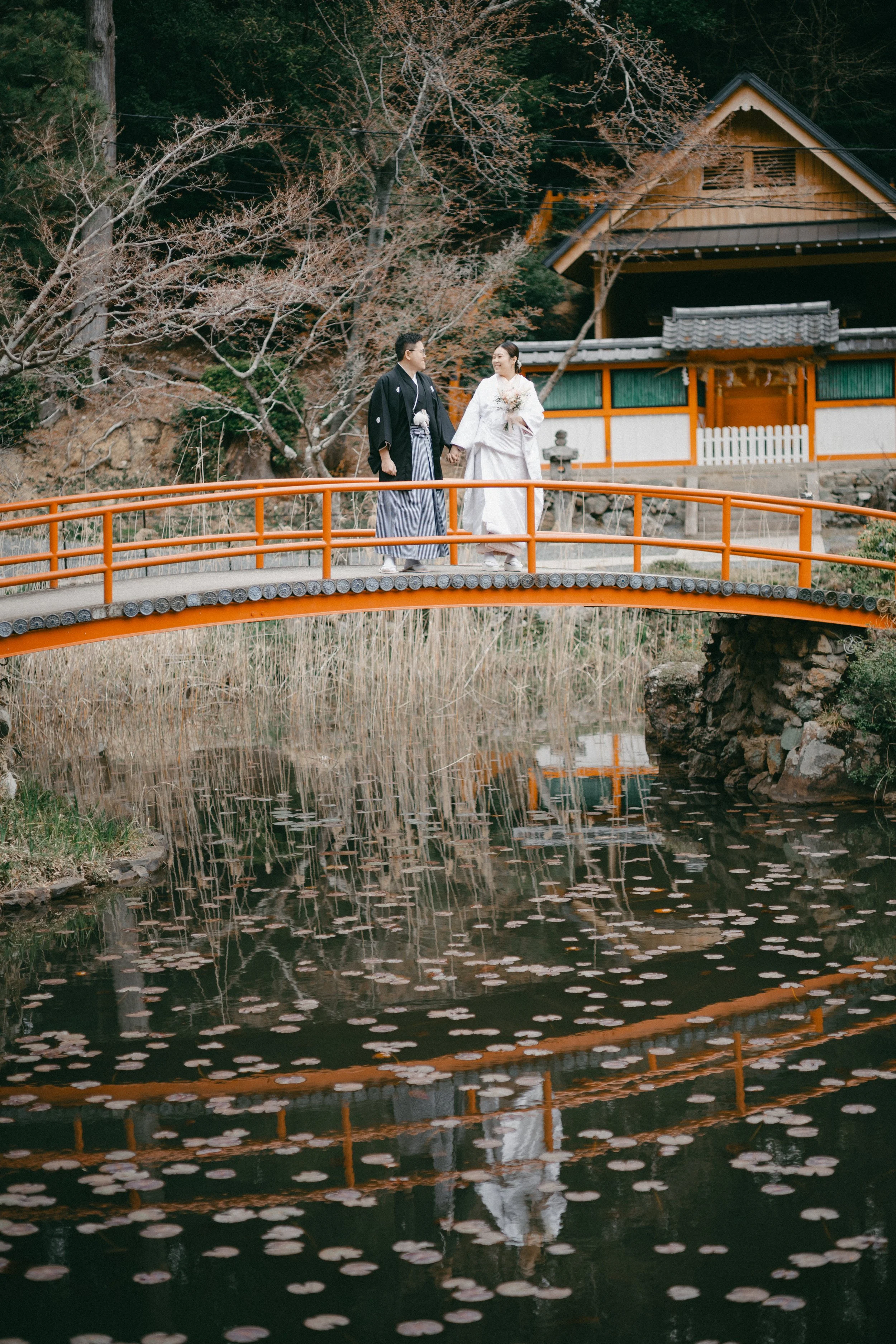 A couple in traditional Japanese wedding attire walking hand in hand on a small orange bridge over a pond, with a traditional Japanese building and bare trees in the background.