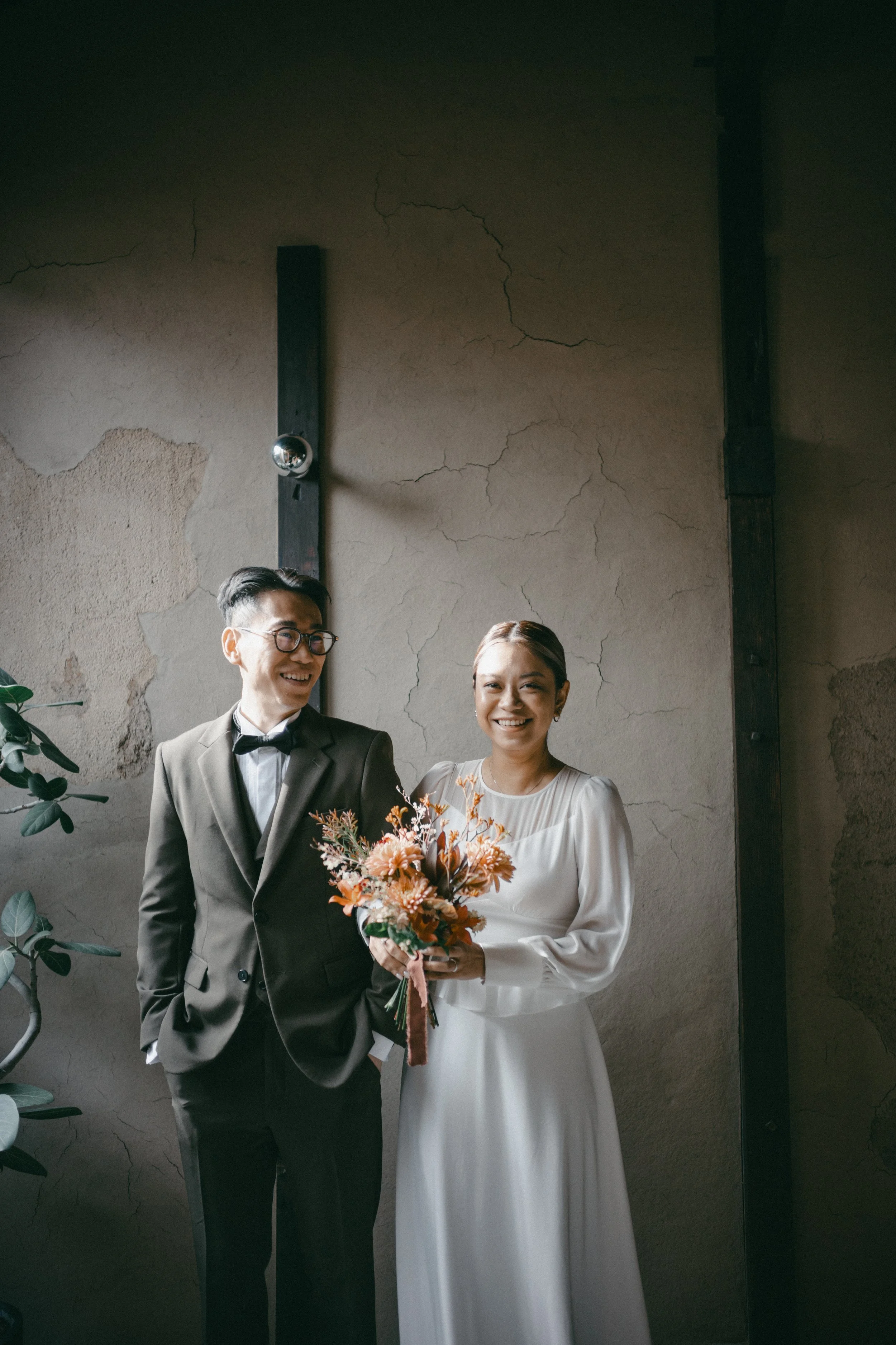 A wedding couple standing together, with the groom in a tuxedo and the bride in a white dress holding a bouquet of flowers, smiling at each other.