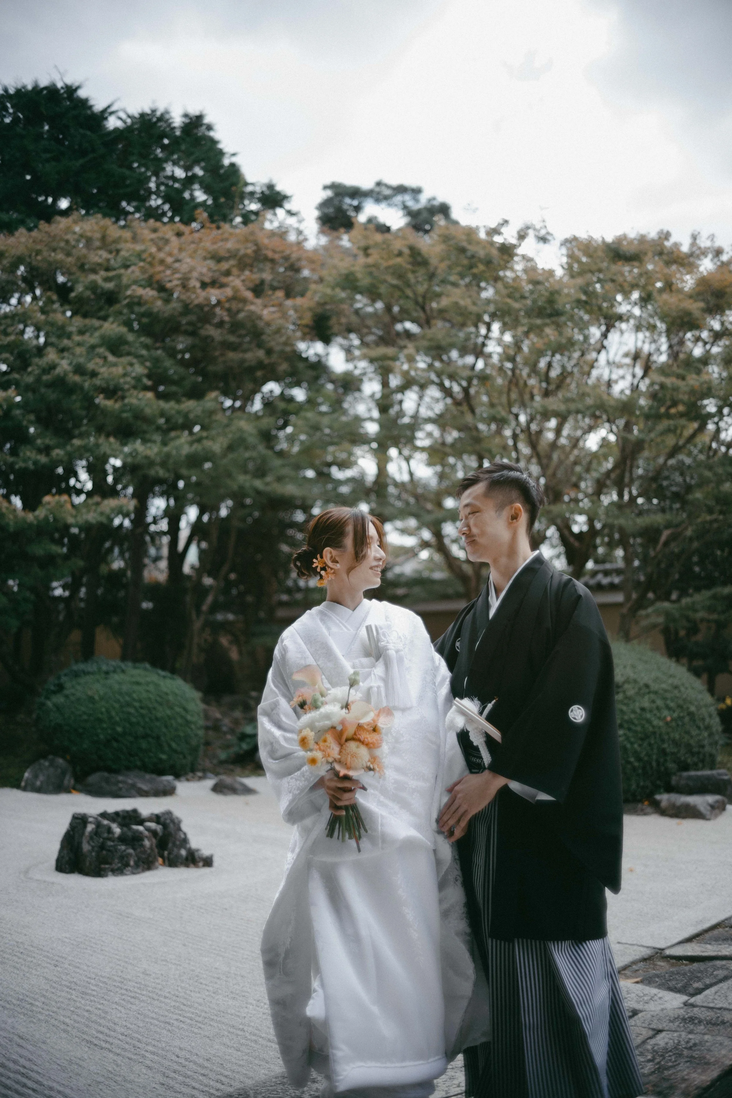 A newlywed Japanese couple dressed in traditional wedding attire, standing outdoors in a garden with trees and rocks, sharing a happy moment.