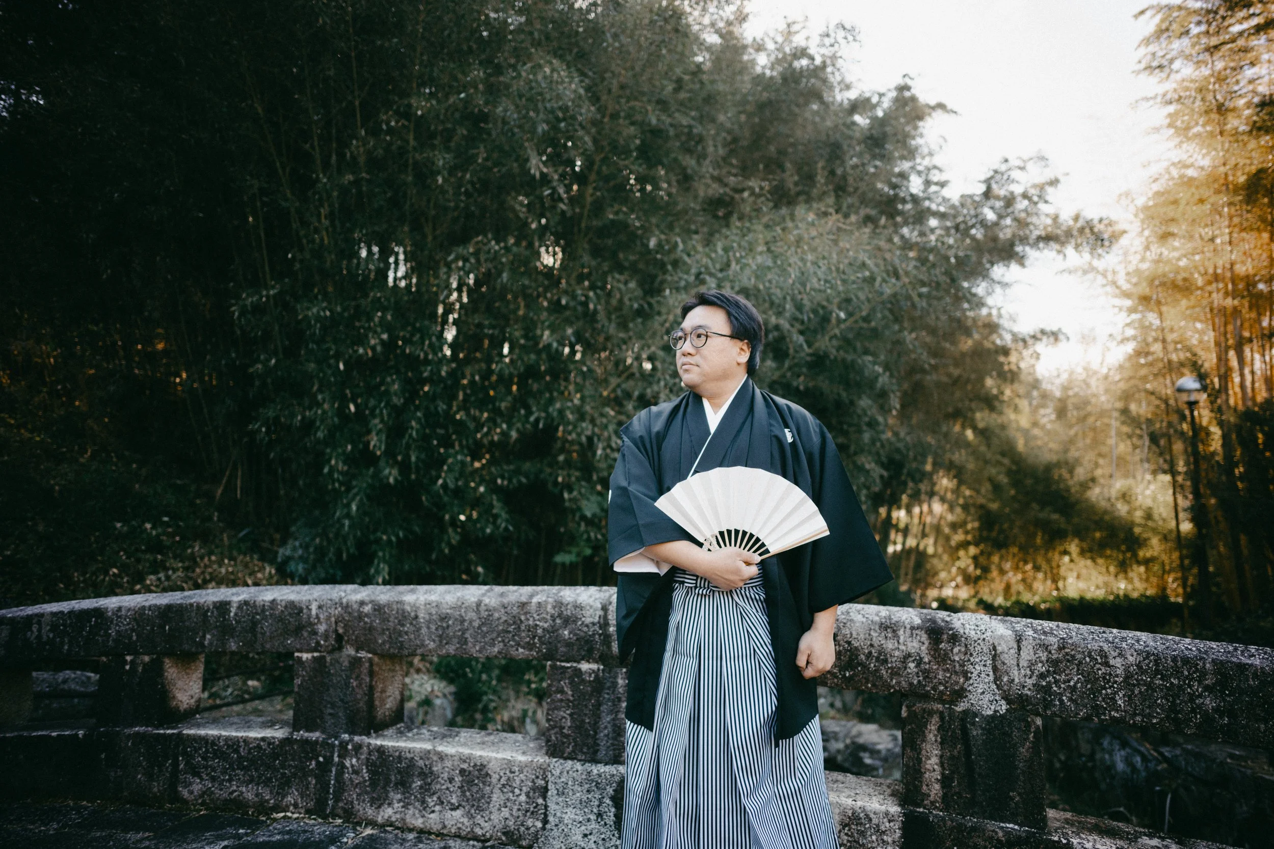 A man wearing traditional Japanese clothing holds a paper fan, standing on a stone bridge with a background of lush trees.