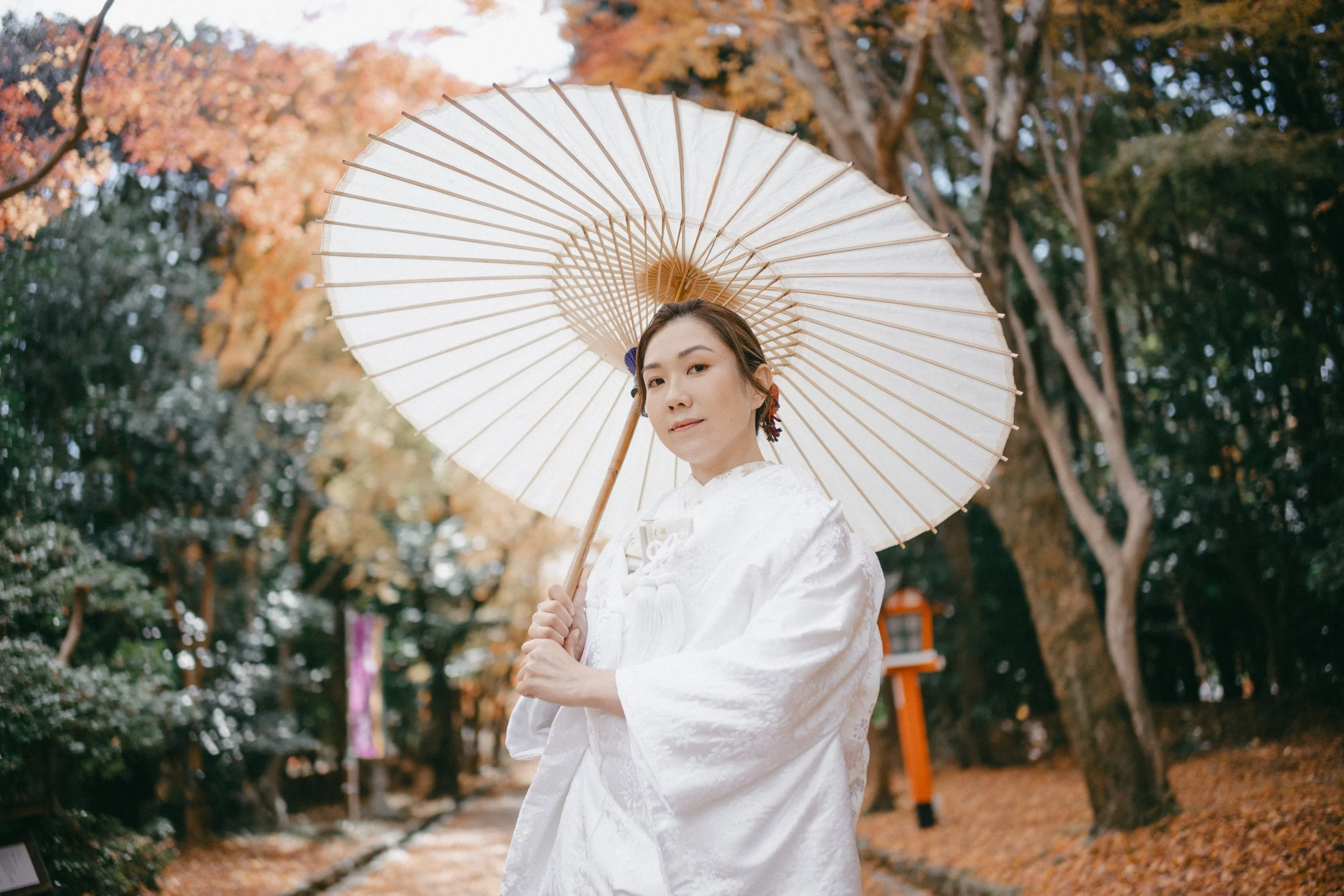 A woman dressed in traditional white Japanese kimono holding a white paper parasol outdoors surrounded by autumn trees.