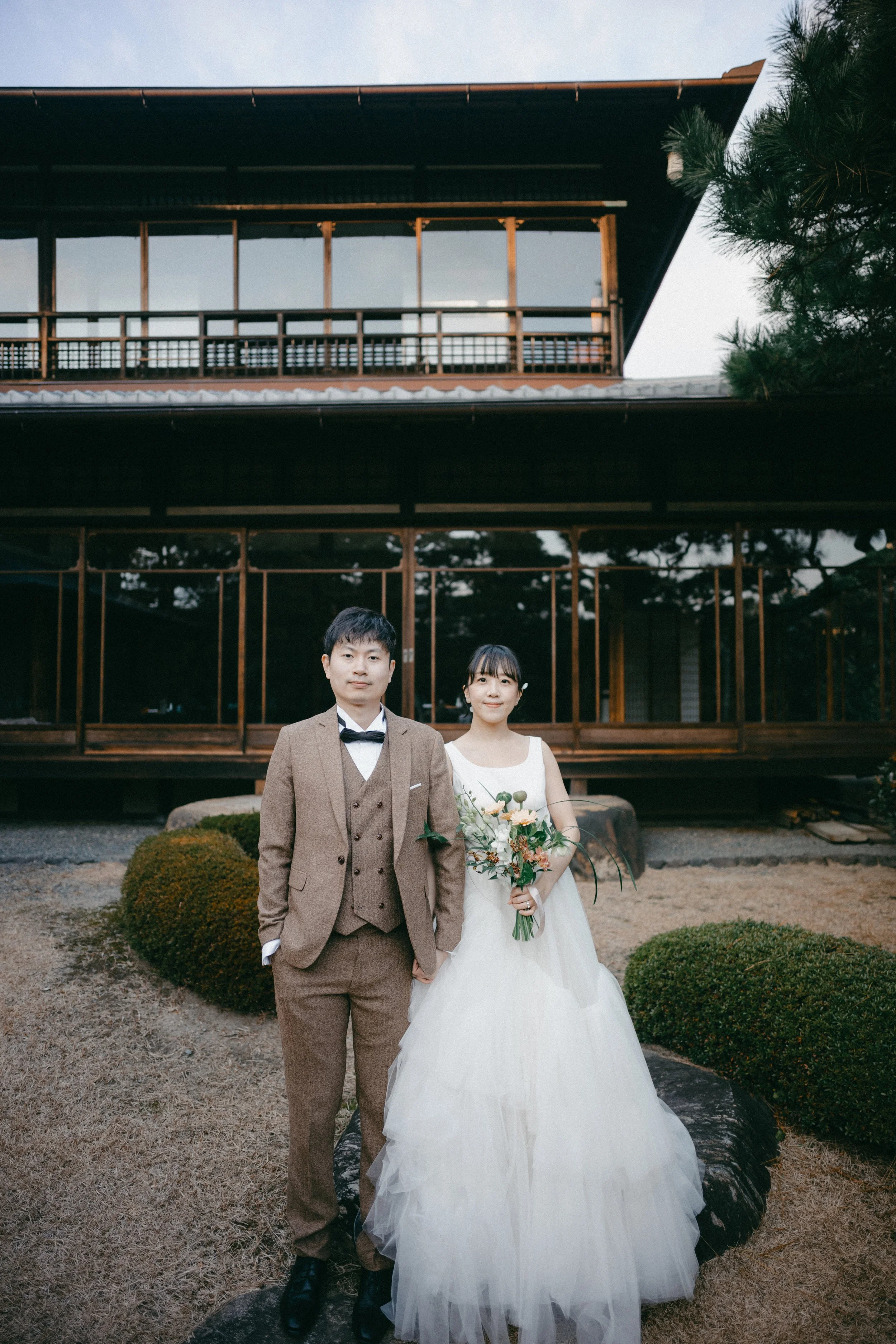 A bride and groom in wedding attire standing outdoors in front of a wooden building, holding hands, with bushes and trees around.