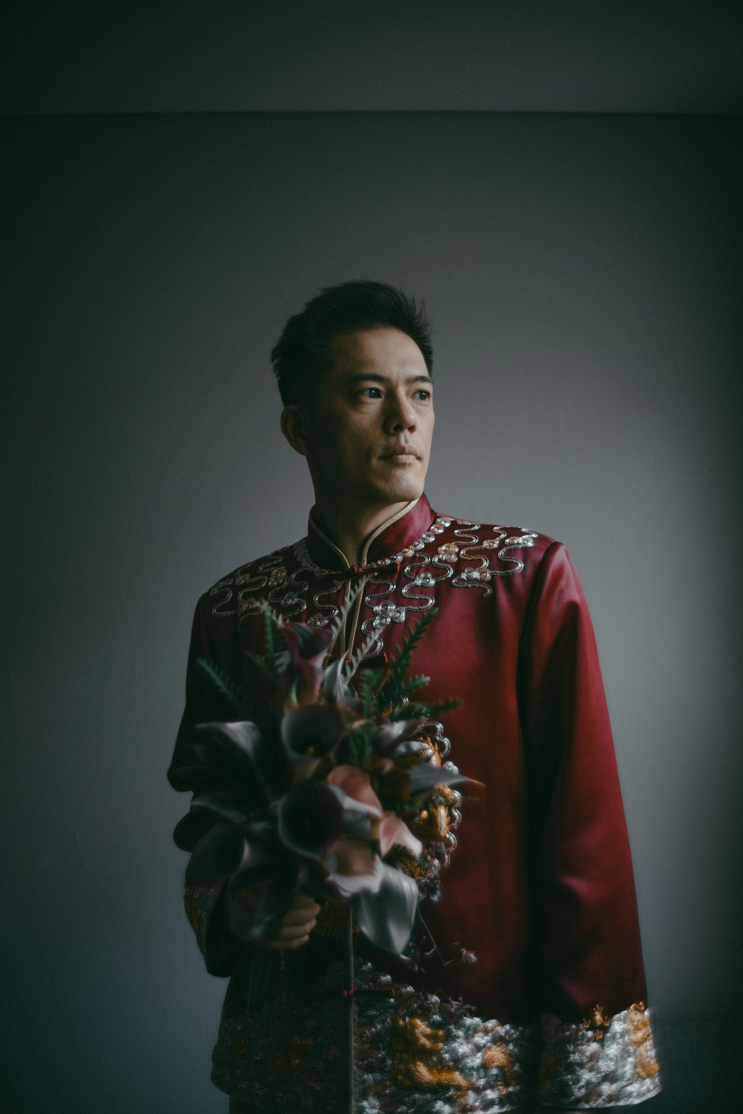 A man in a traditional Chinese red silk jacket with intricate silver embroidery, holding a bouquet of flowers, standing against a plain background.