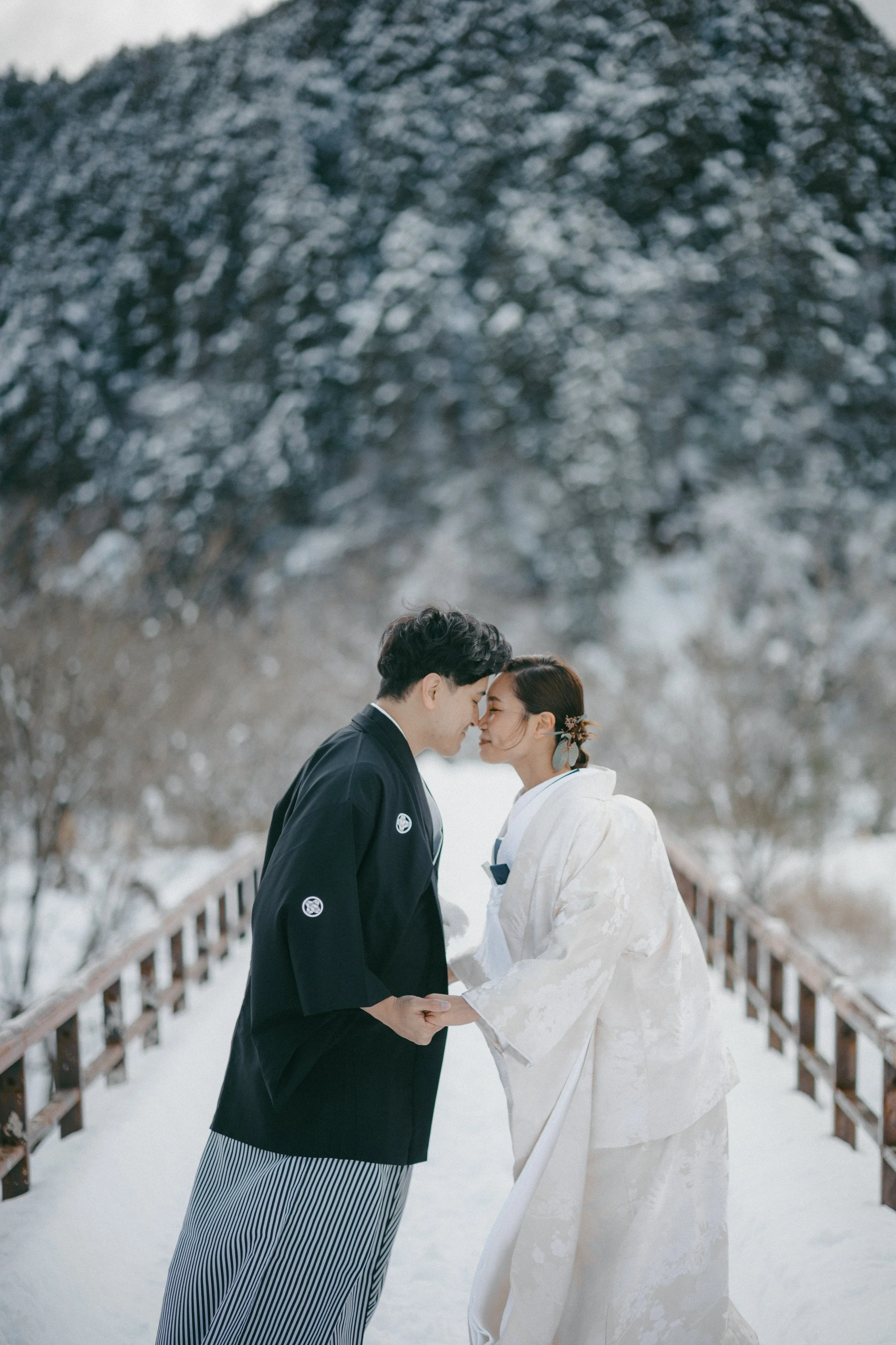 A couple dressed in traditional Japanese wedding attire holding hands and touching foreheads outdoors in a snowy landscape with snow-covered trees in the background.