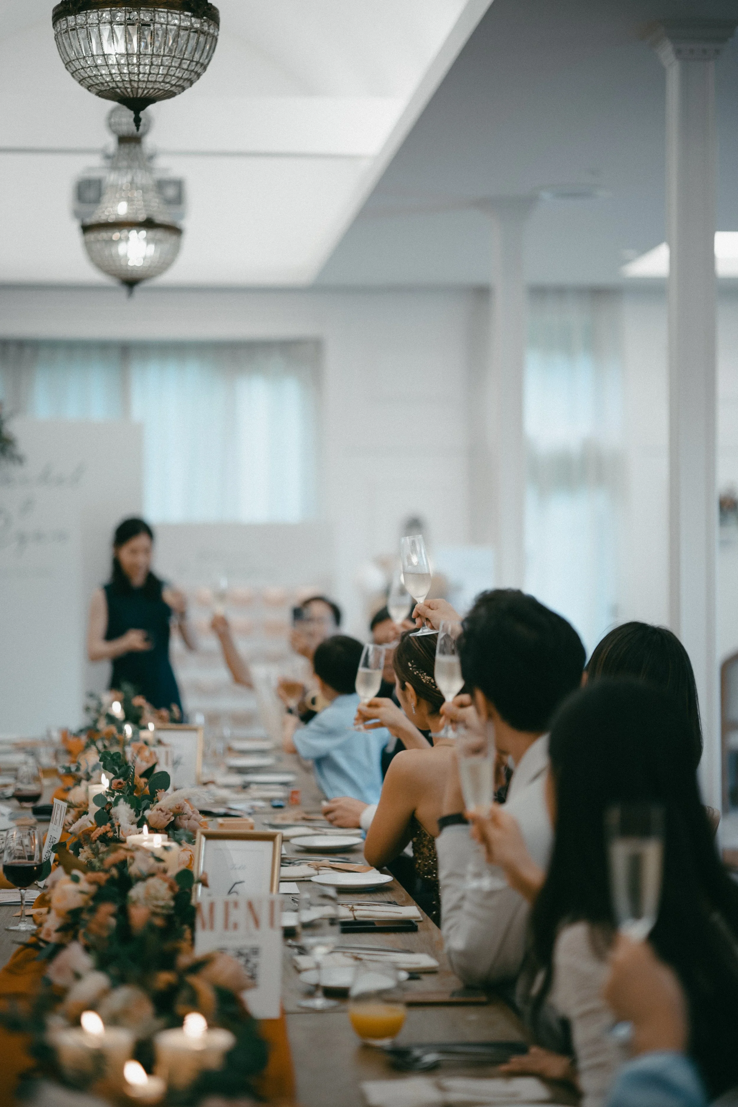 Guests at a wedding reception raising glasses of champagne during a toast, seated at a decorated banquet table with flowers and candles.