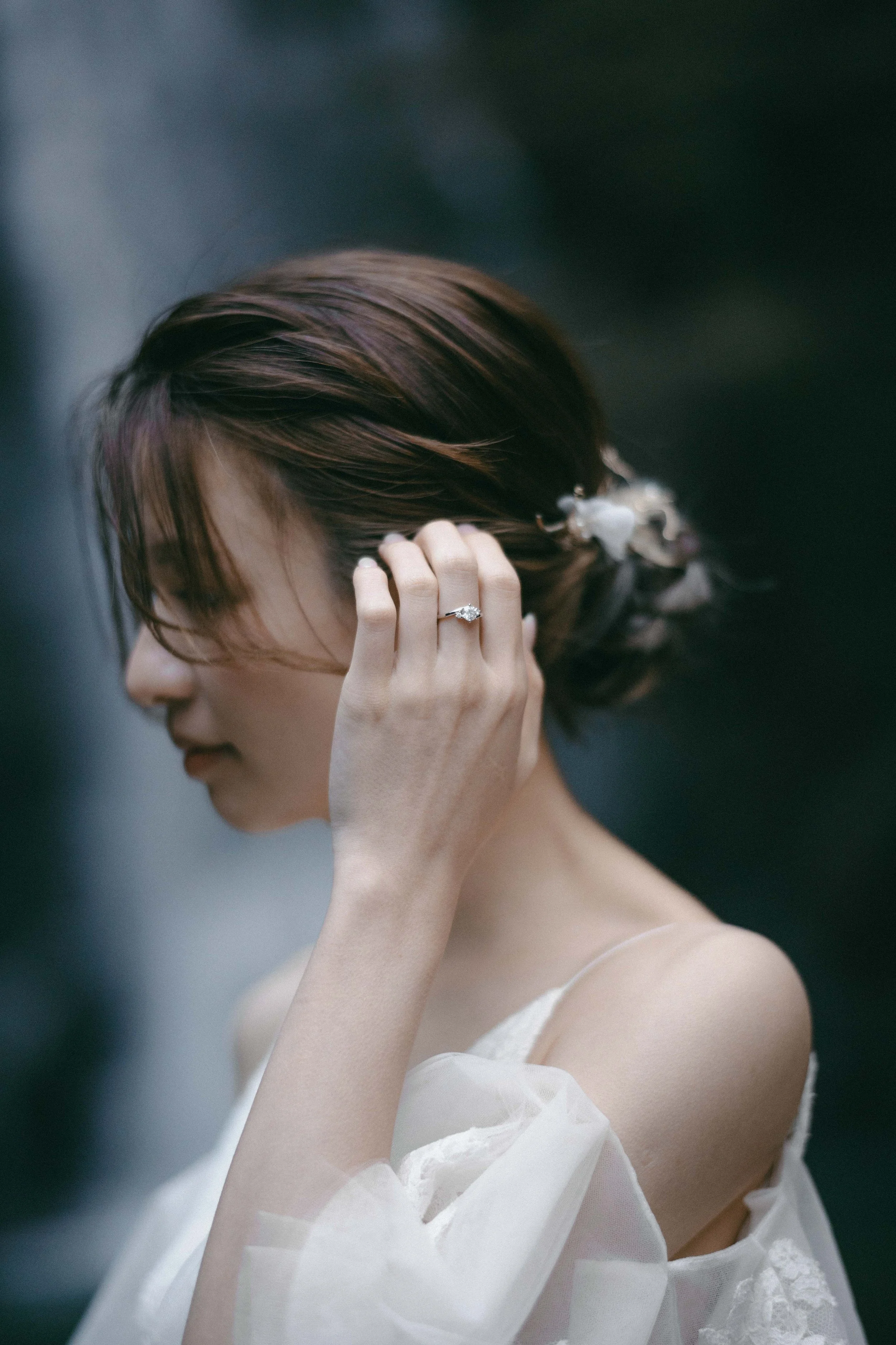 Woman with short brown hair wearing a white dress, adjusting her hair with her left hand, displaying an engagement ring.