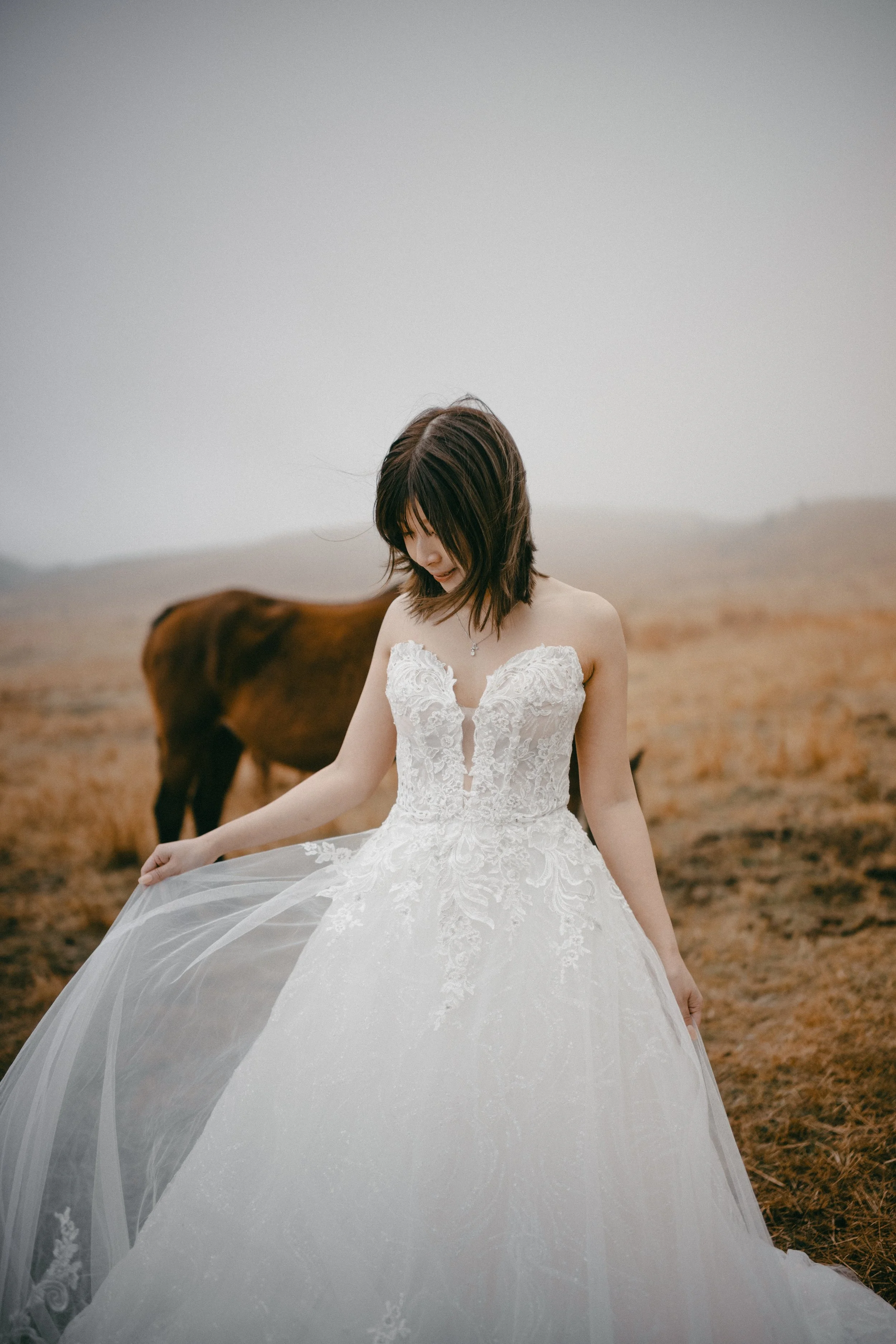 A woman in a white wedding dress standing in a field with a horse in the background.