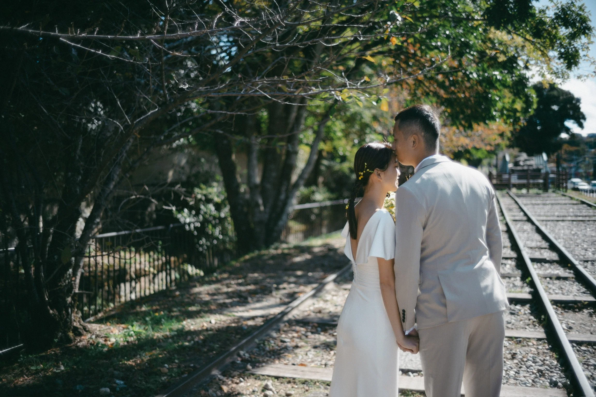 A couple dressed in white holding hands and touching foreheads on train tracks surrounded by trees.