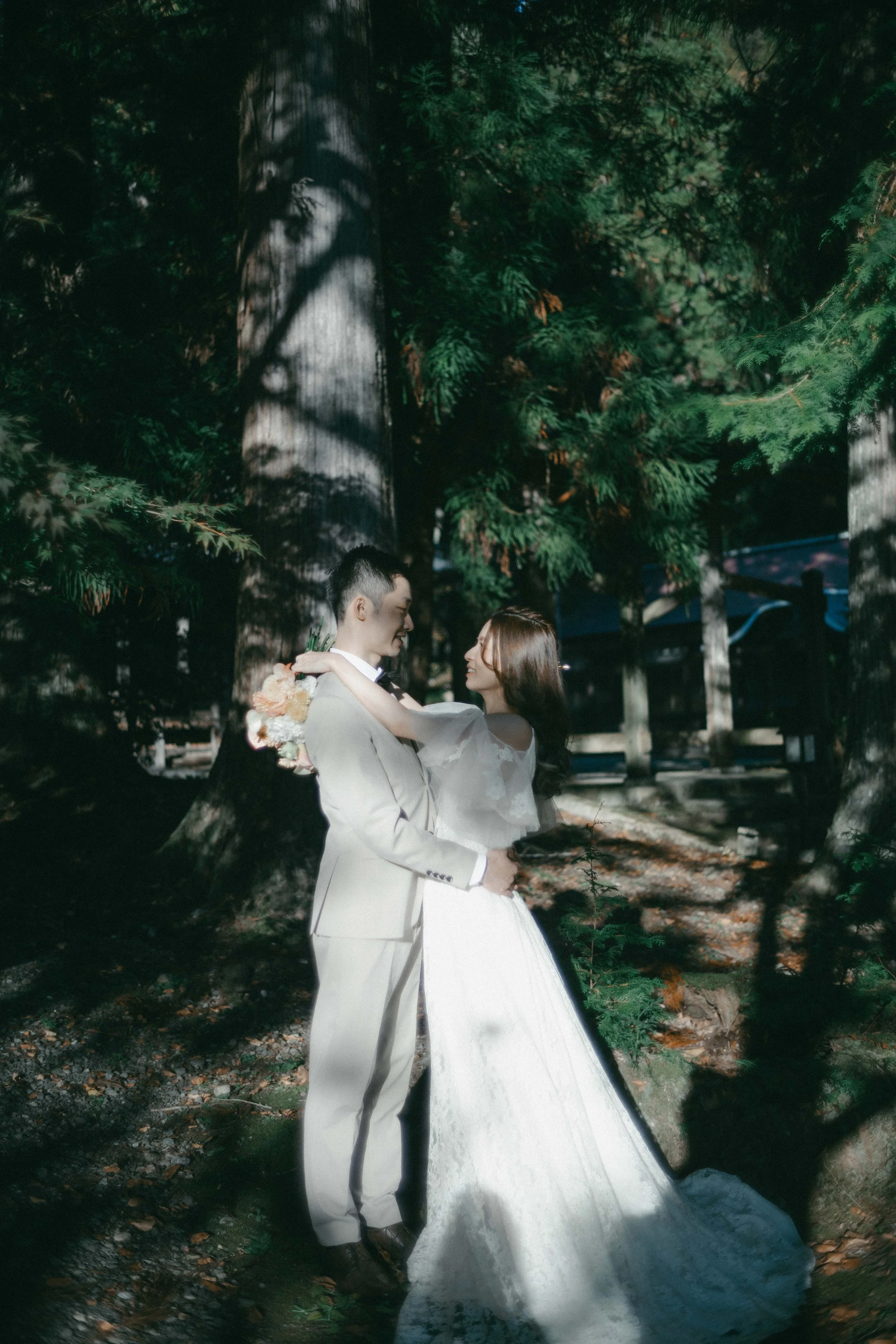 A bride and groom in wedding attire in a forest, embracing each other and gazing into each other's eyes.