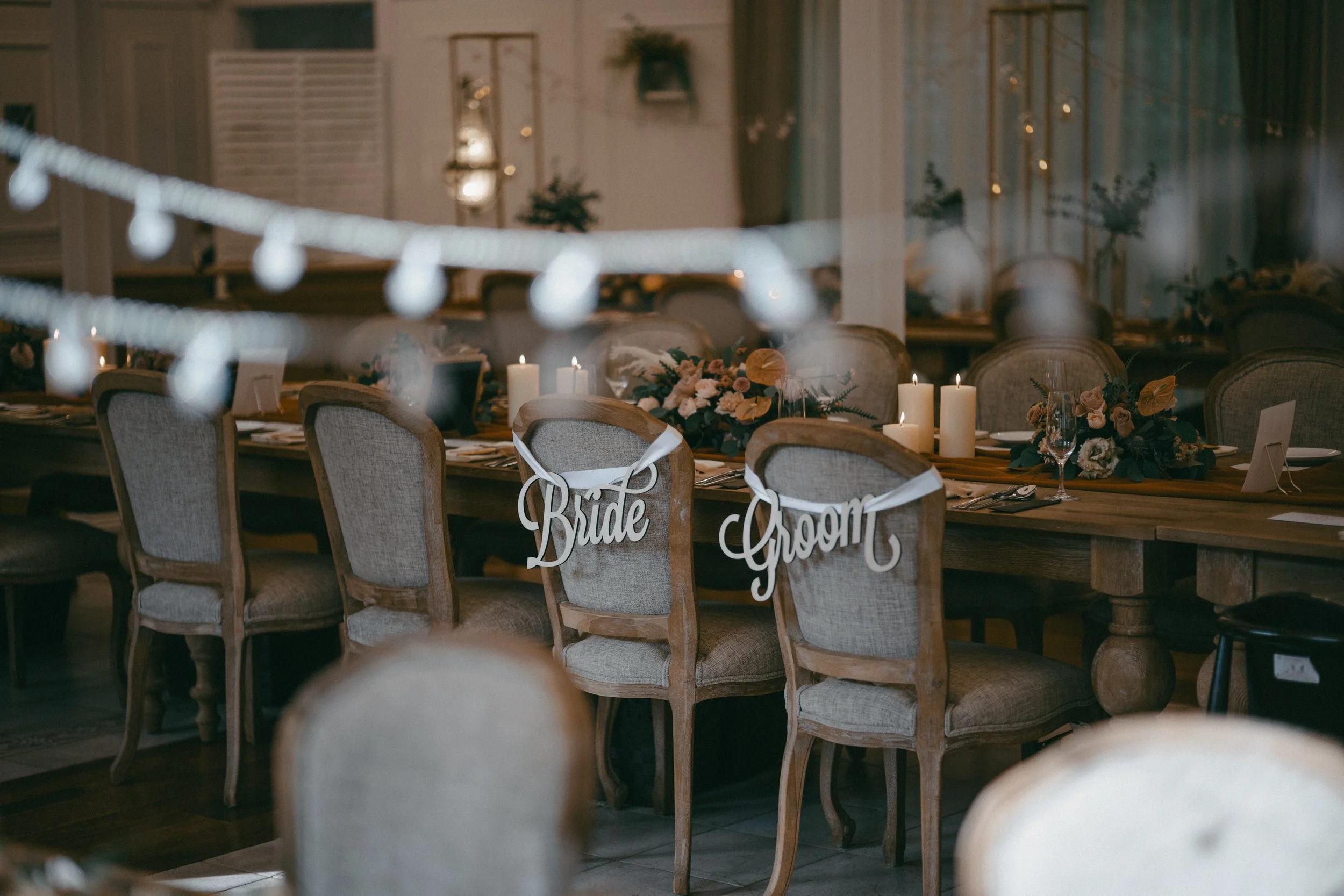 A decorated reception table with a 'Bride' and 'Groom' sign on two chairs, floral centerpieces, lit candles, and place settings, in a warmly lit indoor venue.