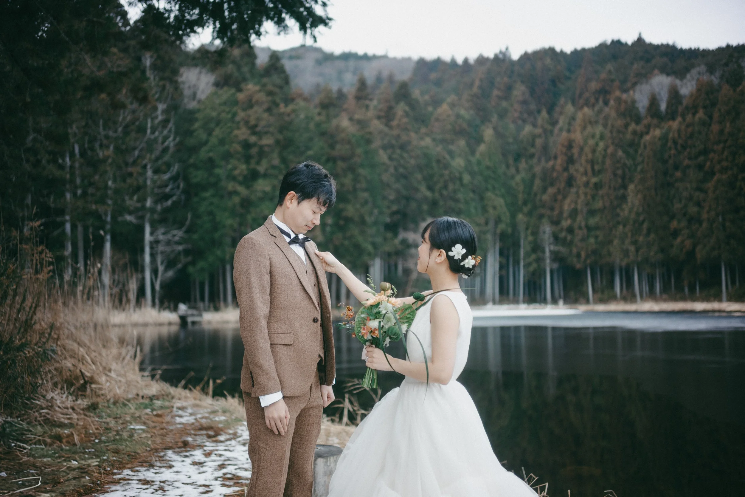 A bride and groom standing by a lake with a forest in the background, the bride touching the groom's chest, holding a bouquet, she has flowers in her hair, and he is dressed in a brown suit.