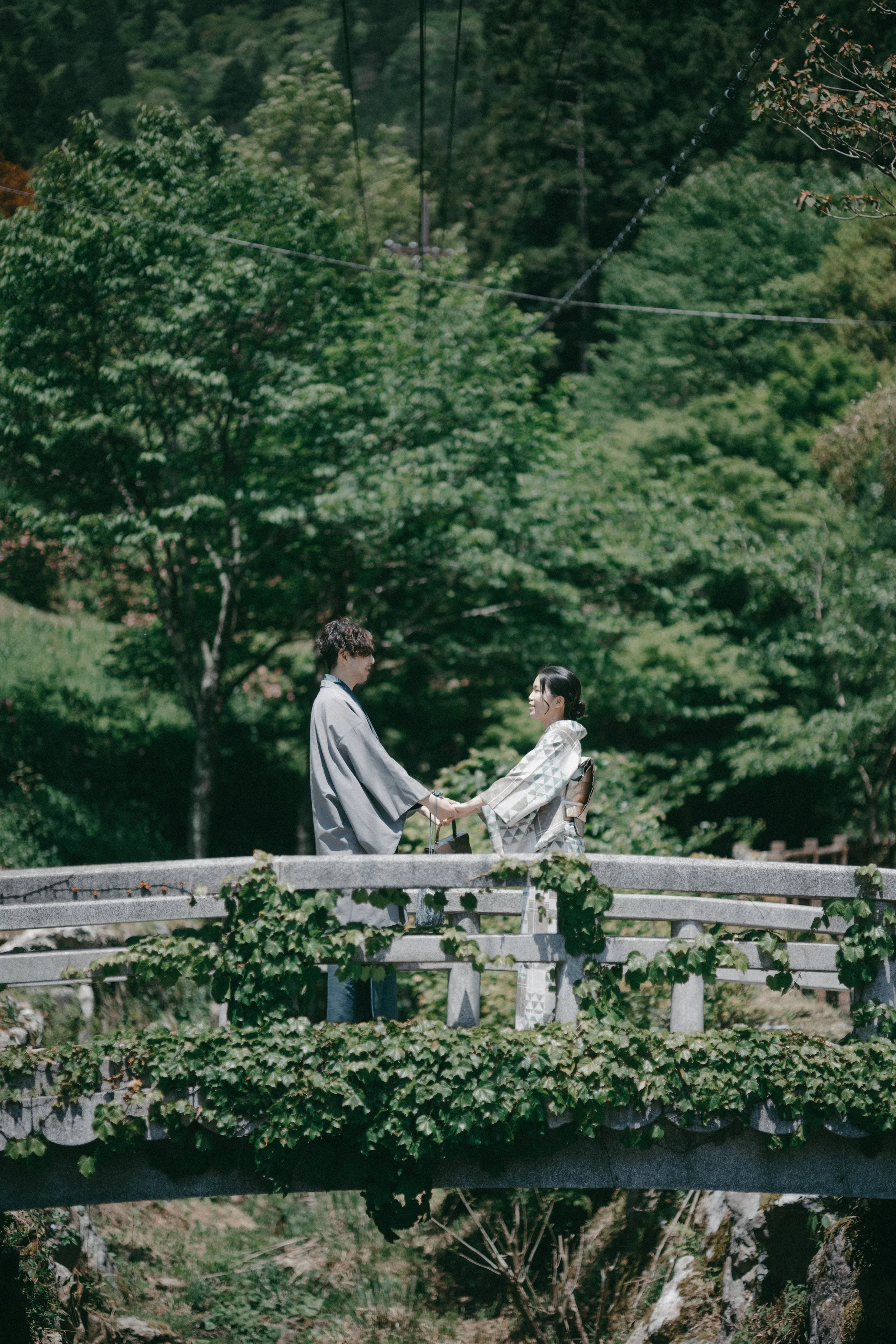A couple in traditional Japanese clothing holding hands on a stone bridge surrounded by lush green trees.