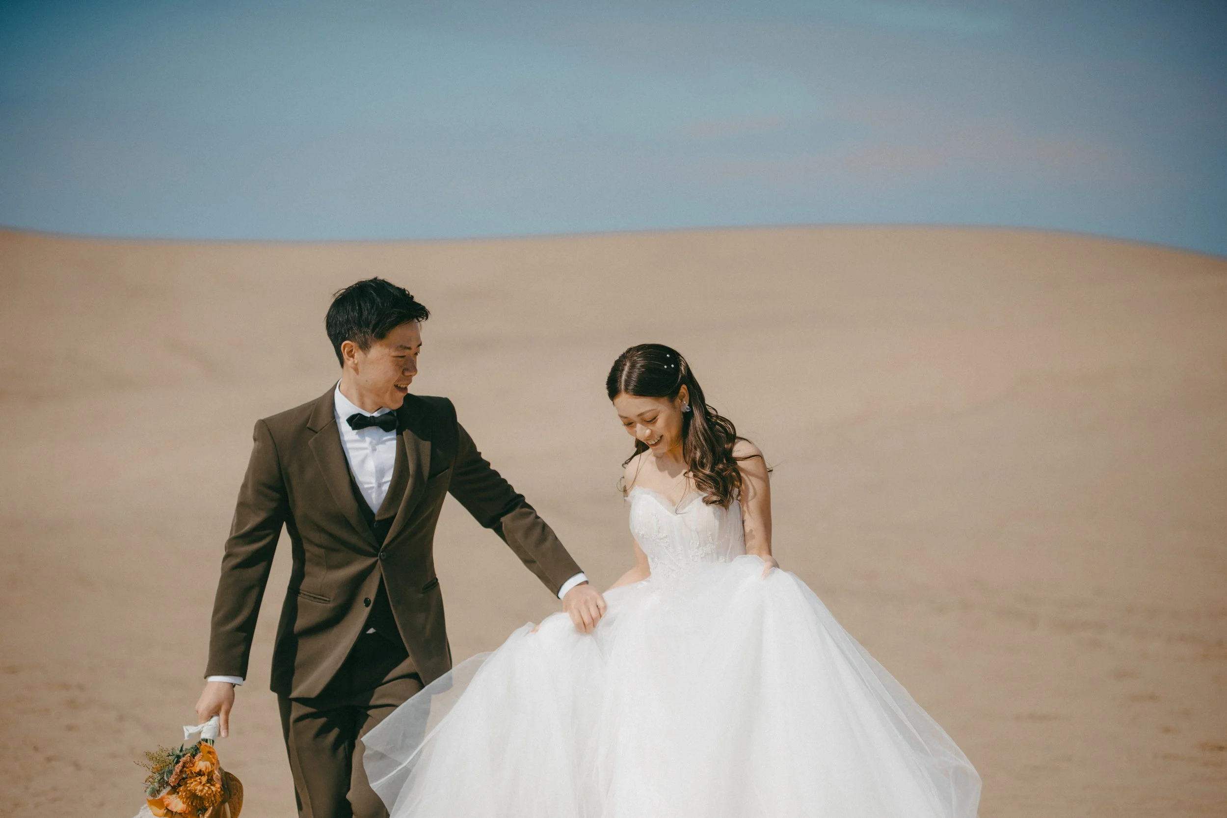 A bride and groom in wedding attire walking on a sandy desert with a blue sky background, smiling and holding hands.