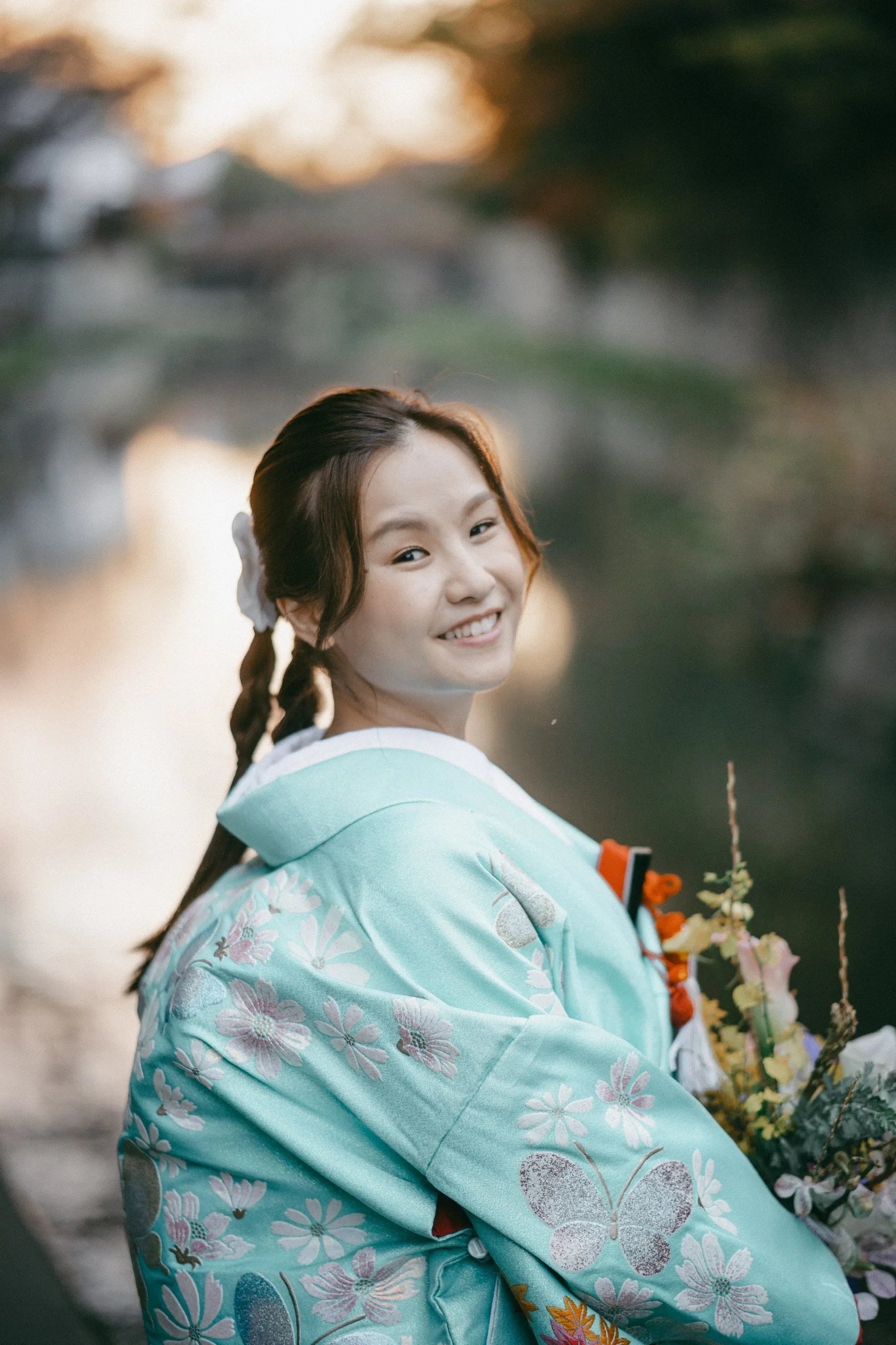 A young woman with a braid tipped with a white flower, wearing a light blue kimono adorned with floral and butterfly patterns, smiles at the camera while holding a bouquet of flowers, with a blurred background of a river and trees at sunset.