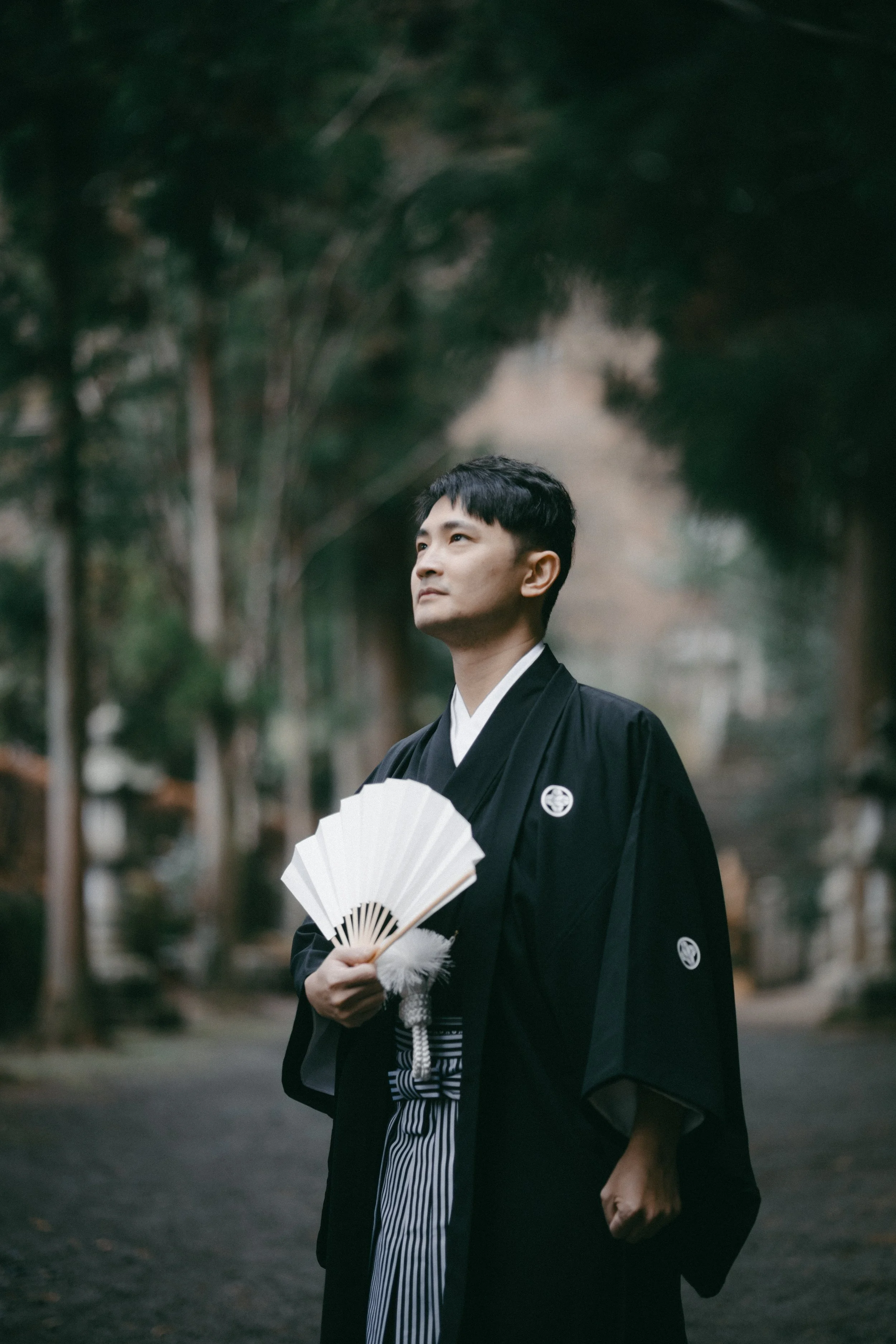 A man dressed in traditional Japanese attire holding a folding fan standing outdoors with trees in the background.