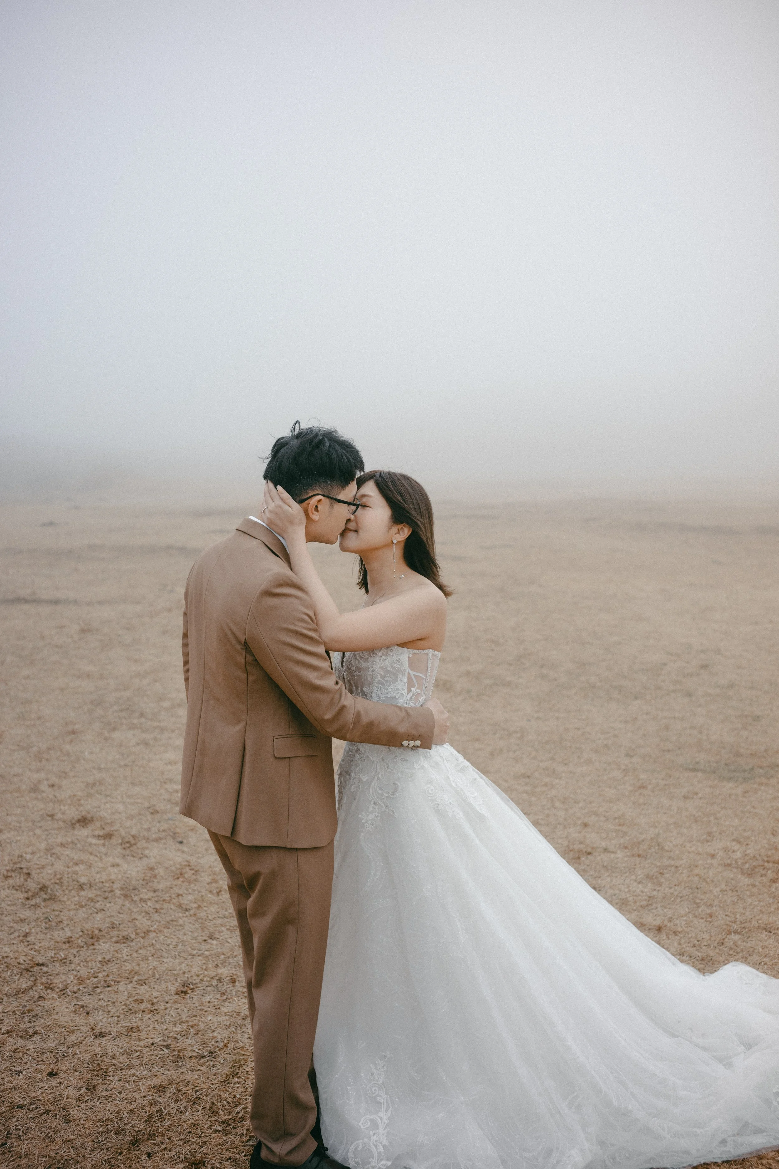 A bride and groom in wedding attire sharing a kiss on a foggy beach