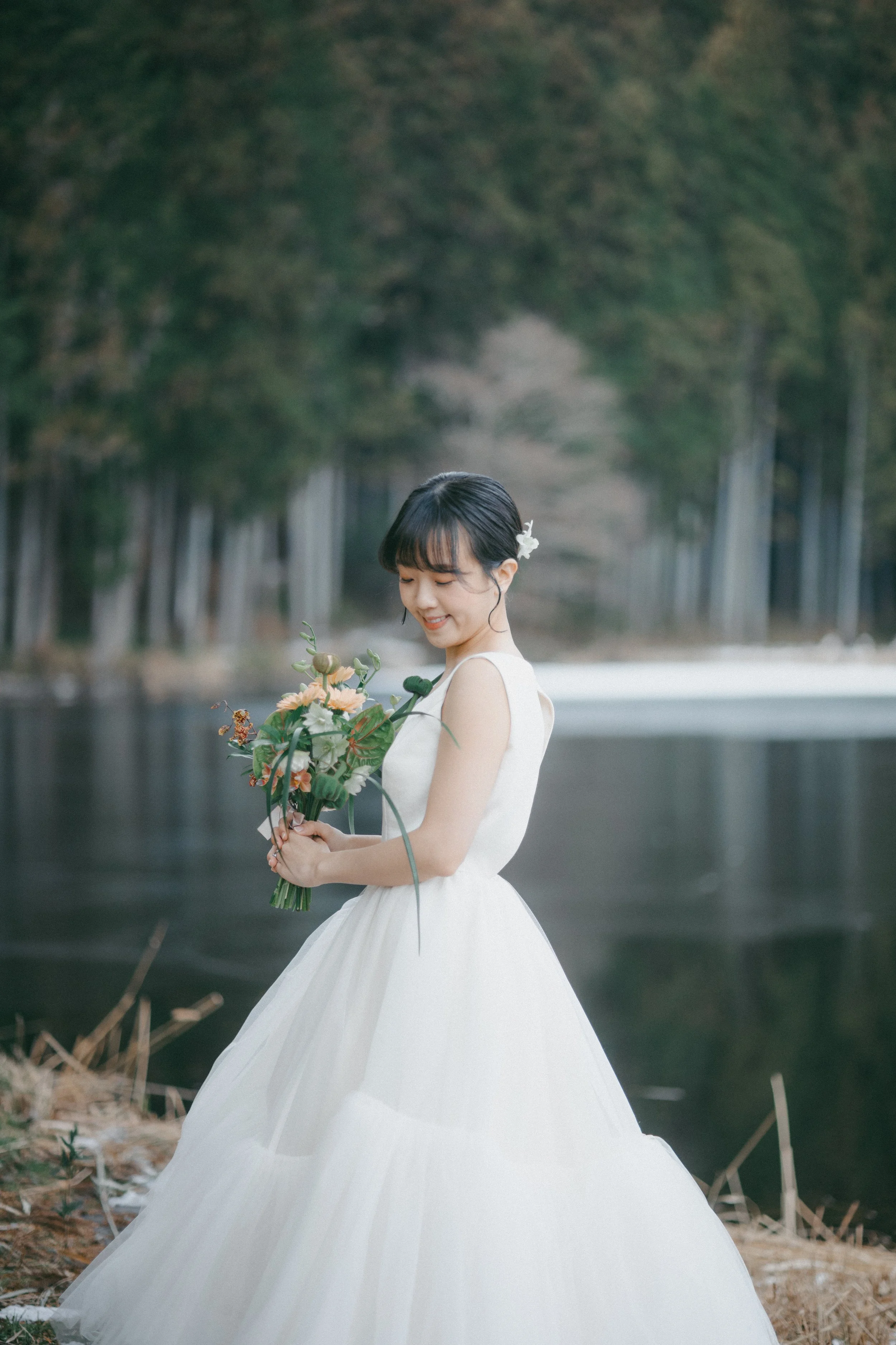 A young woman in a white dress holding a bouquet of flowers by a lake with a forested background.