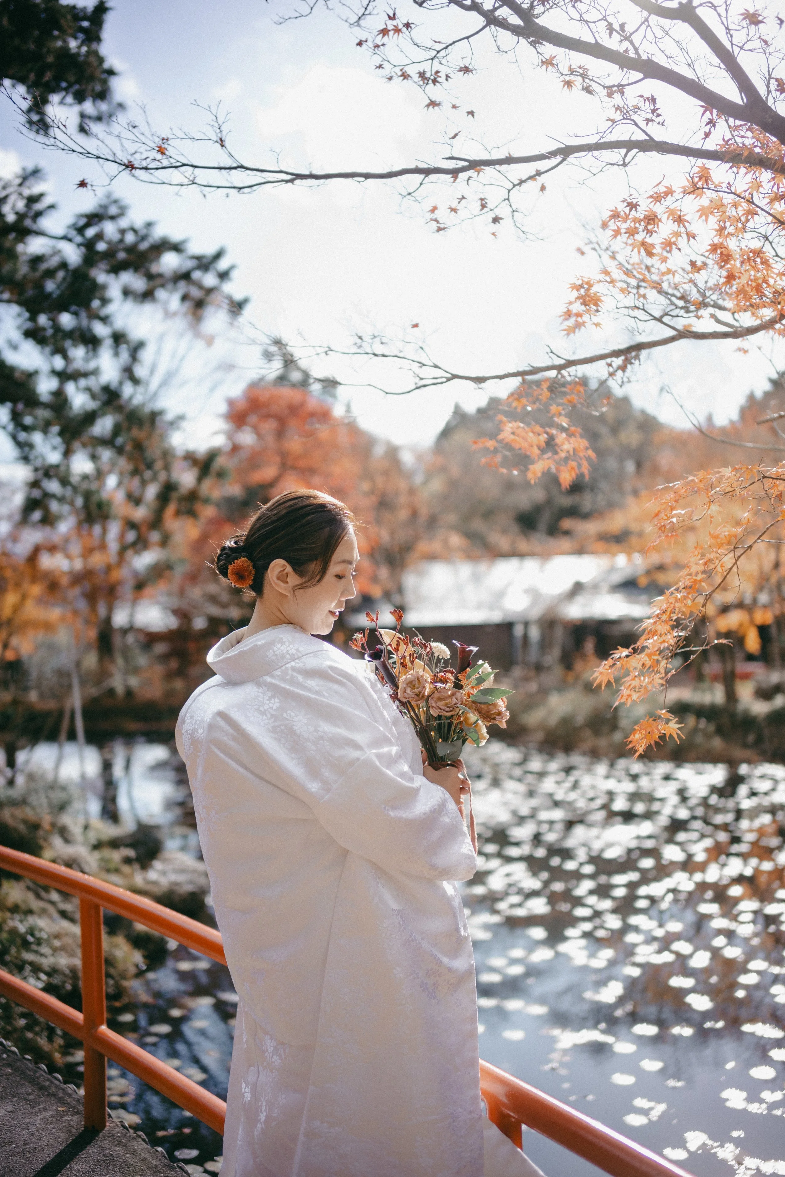 A woman in a white kimono holding a bouquet of flowers, standing on a bridge by a river with autumn trees and fallen leaves in the background.