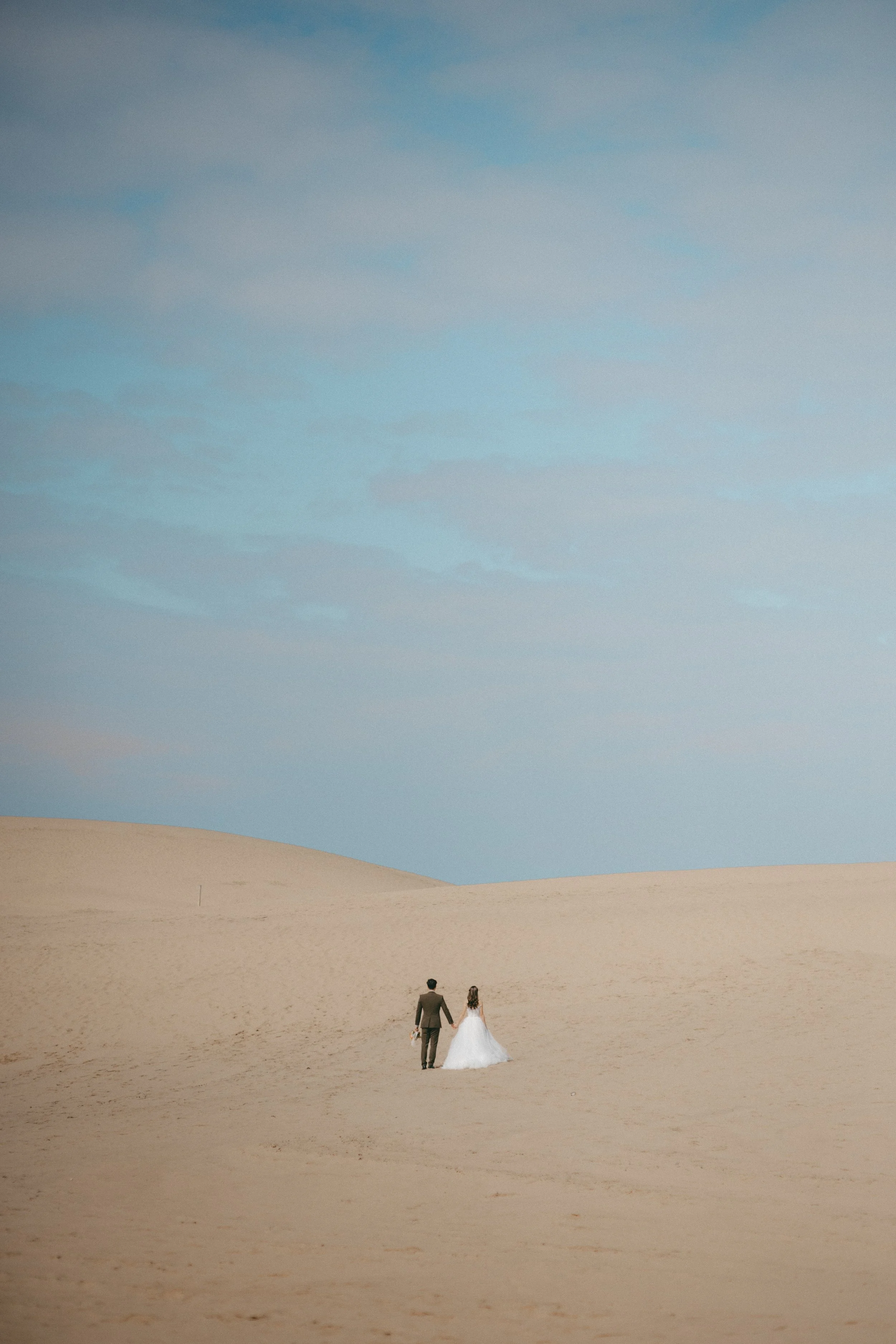 A bride and groom walk hand in hand across a desert landscape with sand dunes under a partly cloudy sky.