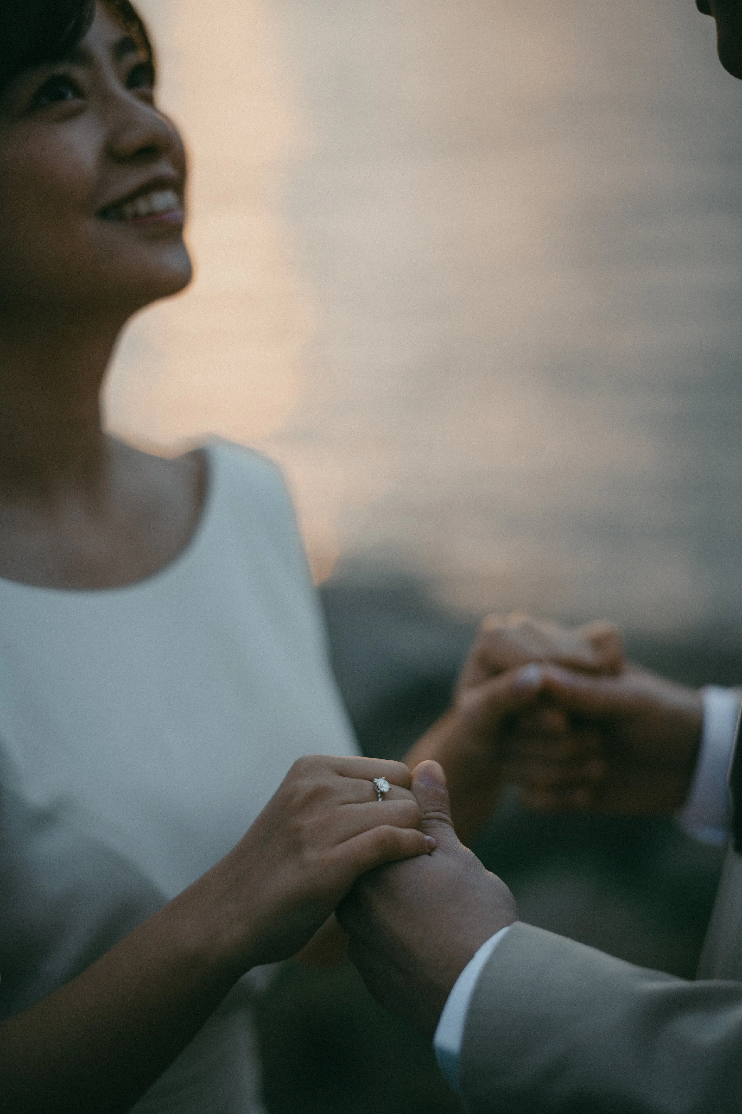 Close-up of a woman and man's hands holding each other, with the woman showing a wedding ring, against a blurred natural background.