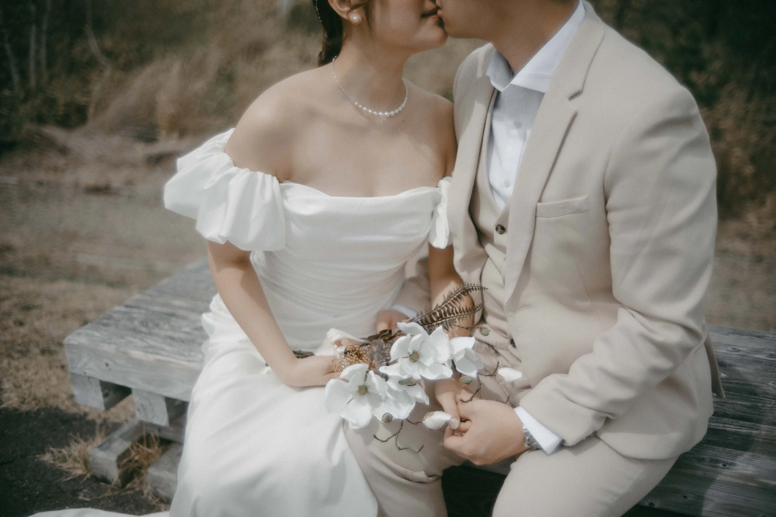A bride and groom sitting on a wooden bench, close together, with the couple kissing. The bride is wearing an off-shoulder white dress and pearl necklace, and the groom is in a beige suit with a white shirt. The bride is holding a bouquet with white 