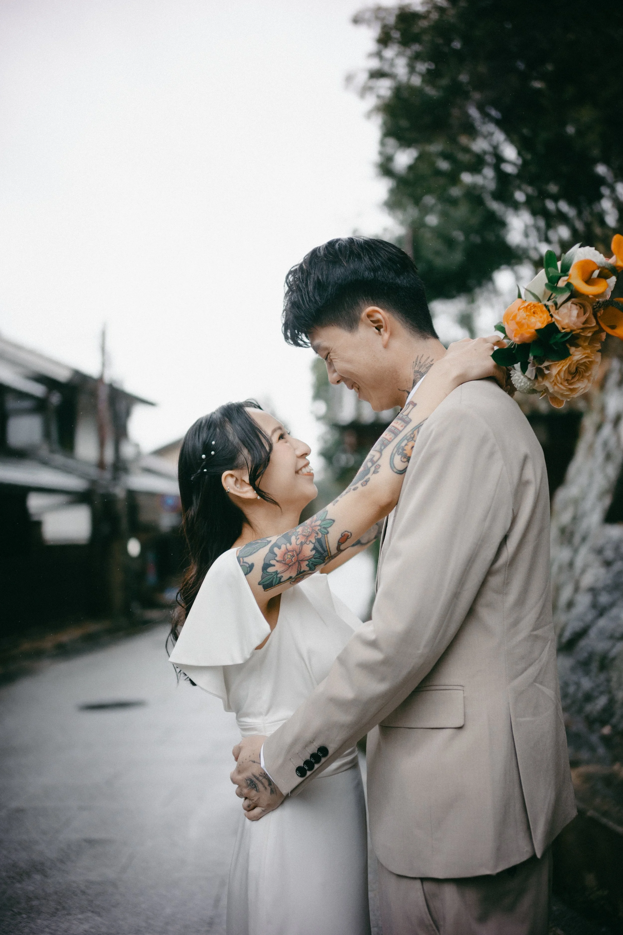 A couple smiling and looking into each other's eyes, embracing outdoors with trees and buildings in the background.