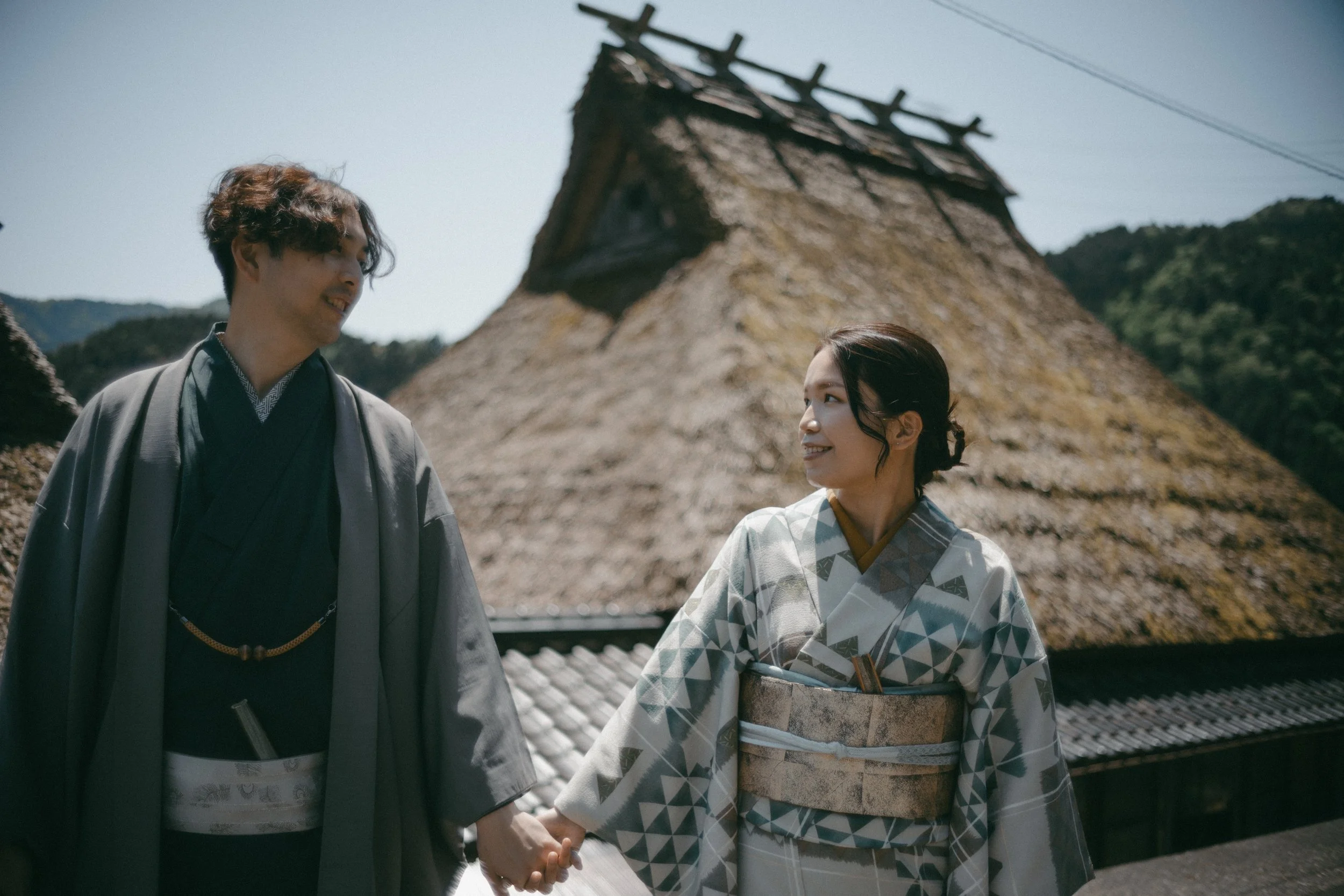 A young Japanese couple dressed in traditional kimonos, holding hands and smiling at each other, with a thatched roof building in the background and mountains under a clear sky.