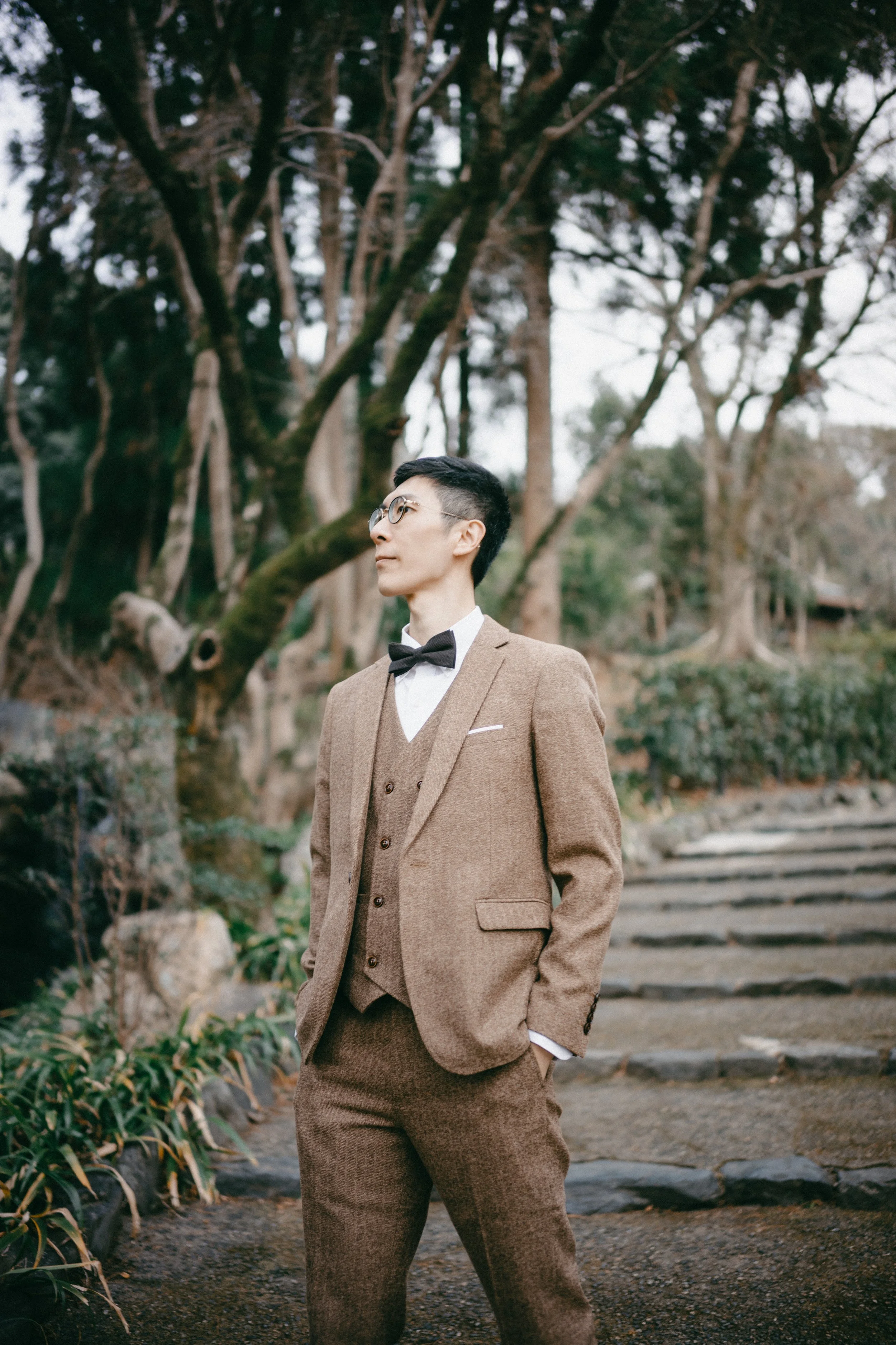 A man dressed in a brown suit with a bow tie, white shirt, and glasses standing outdoors on a stone path surrounded by trees and greenery.
