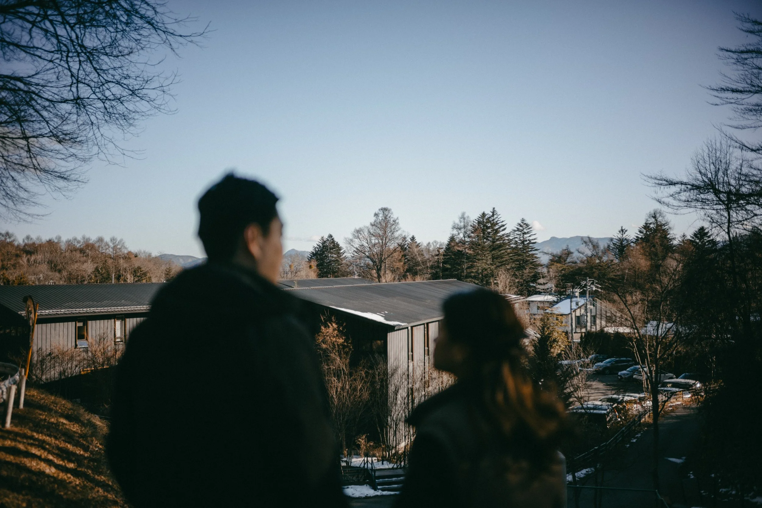 Blurred silhouettes of a man and woman standing outdoors during daylight with trees, buildings, and a parking lot in the background.