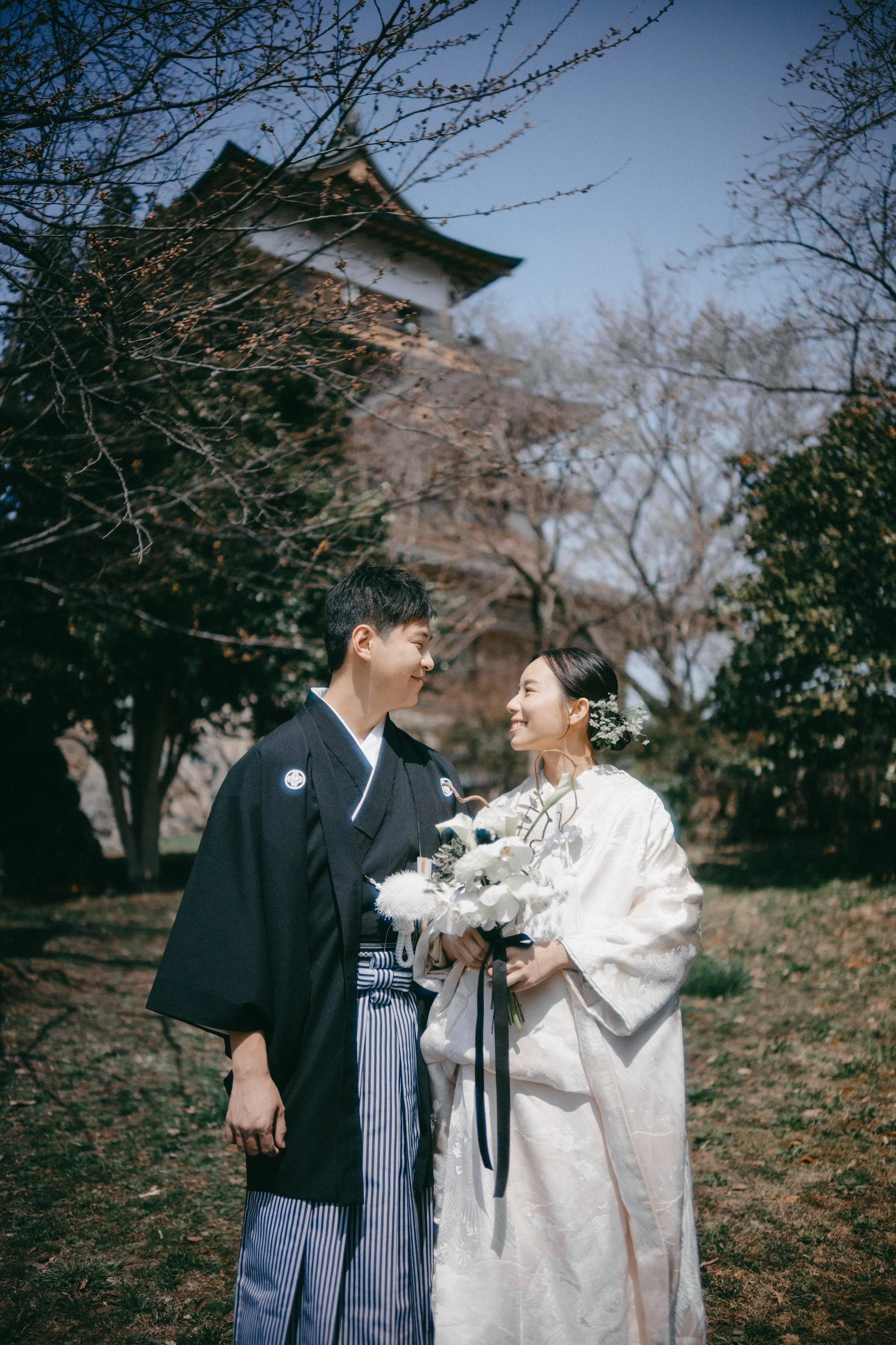 A couple in traditional Japanese wedding attire standing outdoors in front of a temple and leafless trees, smiling at each other, with the woman holding a bouquet of white flowers.