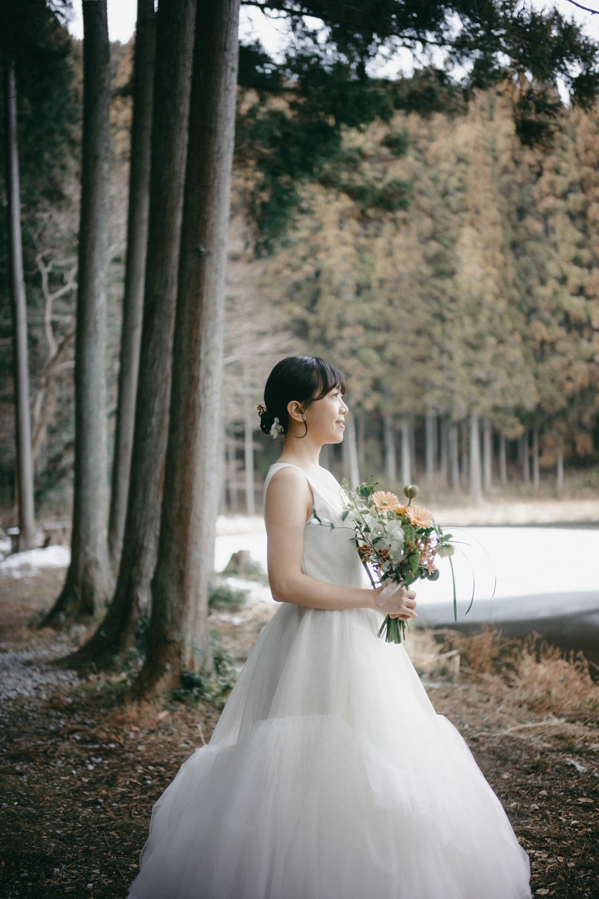 A woman in a white wedding dress holding a bouquet of flowers in a forest clearing.