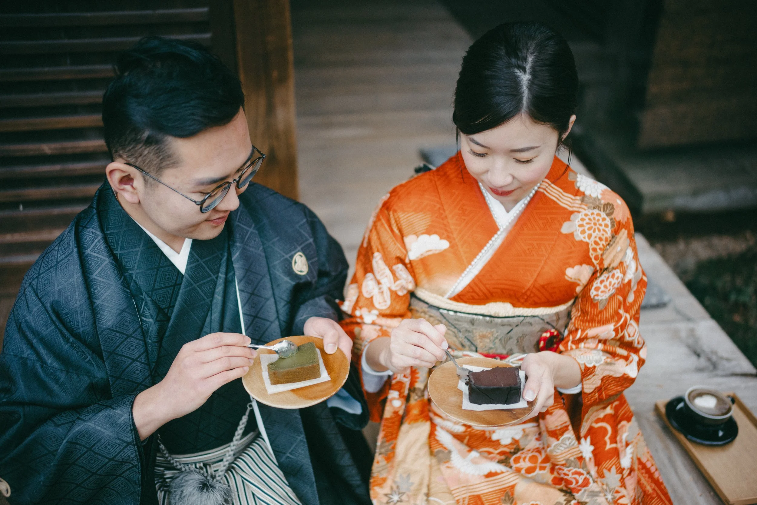 A man and a woman wearing traditional Japanese kimonos sit outdoors, enjoying desserts on small wooden plates.