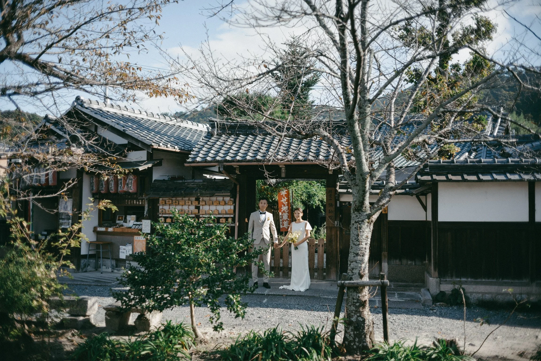 A newlywed couple holding hands standing outside a traditional Japanese building with a tiled roof, surrounded by garden plants and trees.