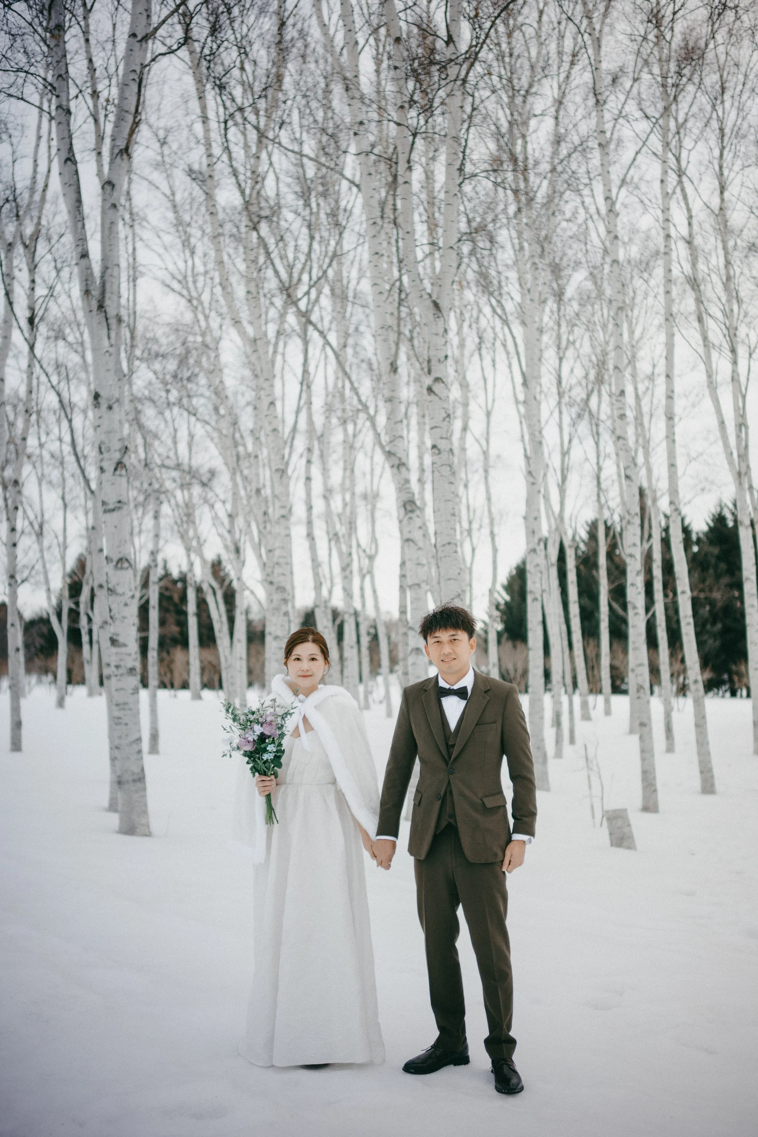 A bride and groom holding hands in a snowy forest. The bride is wearing a white dress with a white cape and holding a bouquet, while the groom is dressed in a brown suit with a black bow tie.