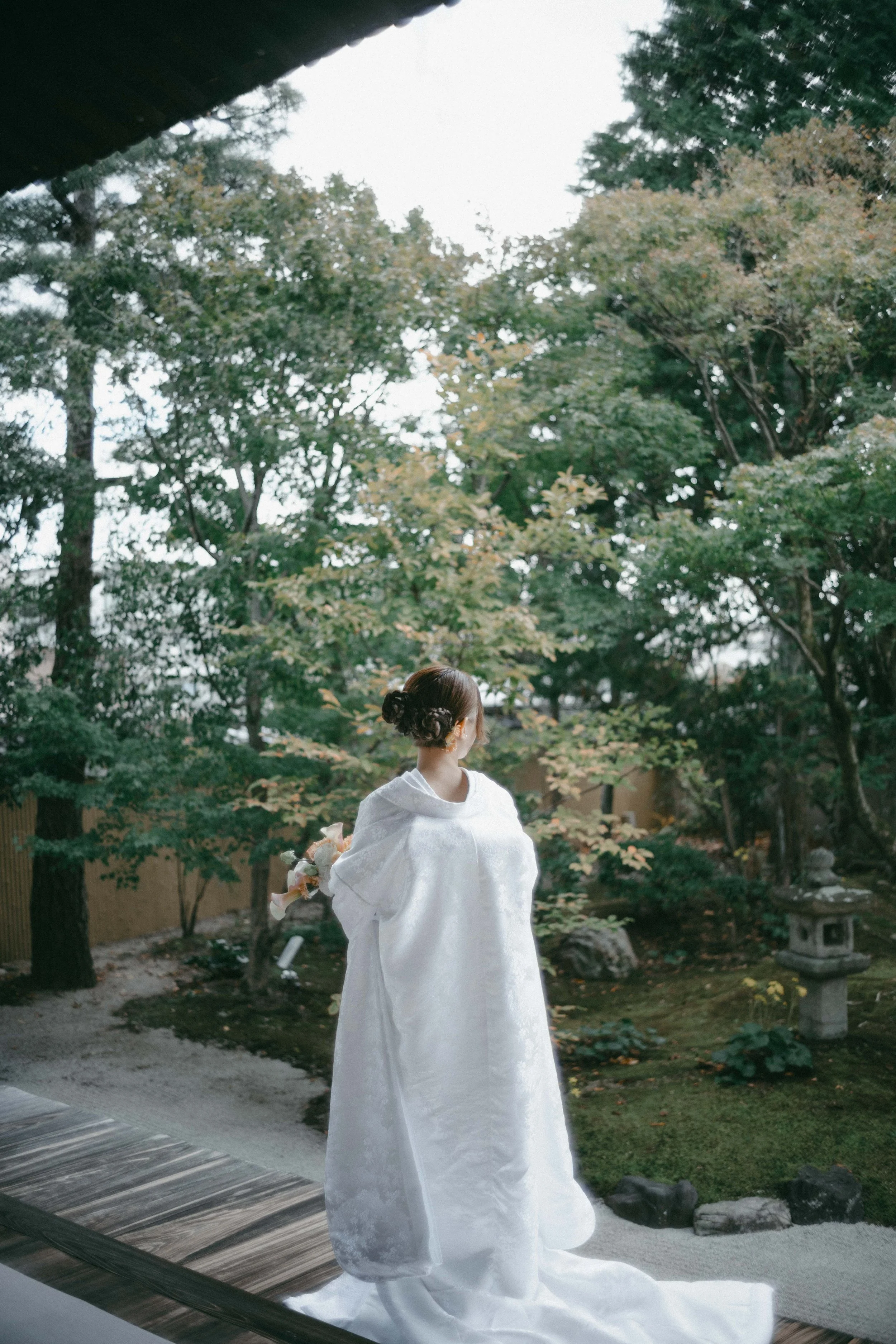 A woman in a white wedding gown standing in a traditional Japanese garden with lush trees, a stone lantern, and gravel ground, viewed from the back.