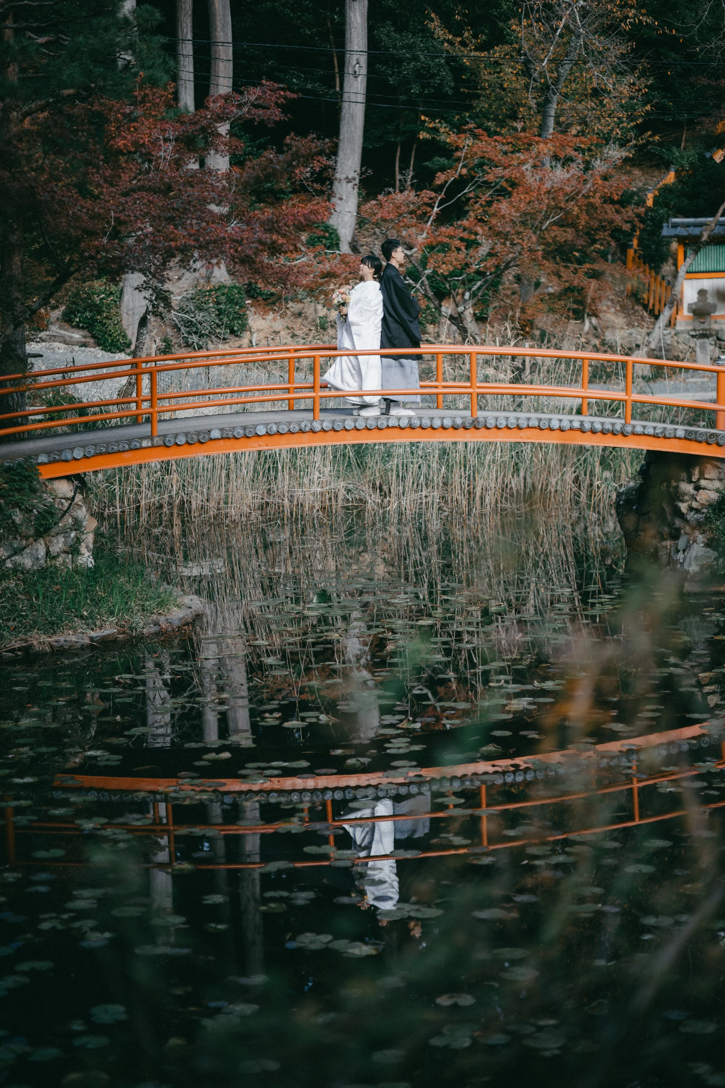A bride and groom in traditional Japanese wedding attire walking across a small orange bridge over a pond with lily pads, surrounded by autumn foliage and trees.
