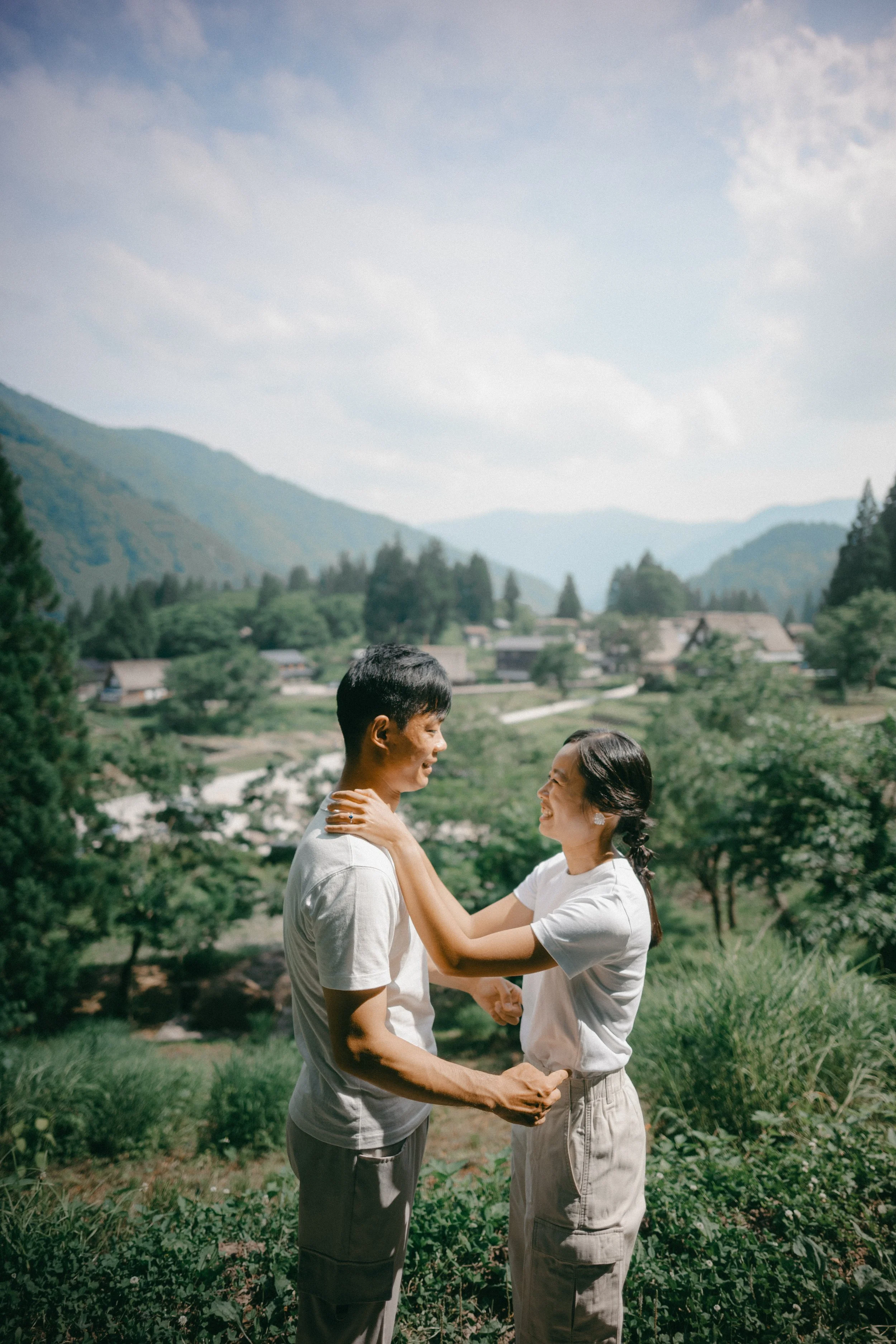 A young man and woman smiling at each other outdoors in a green landscape with mountains in the background.