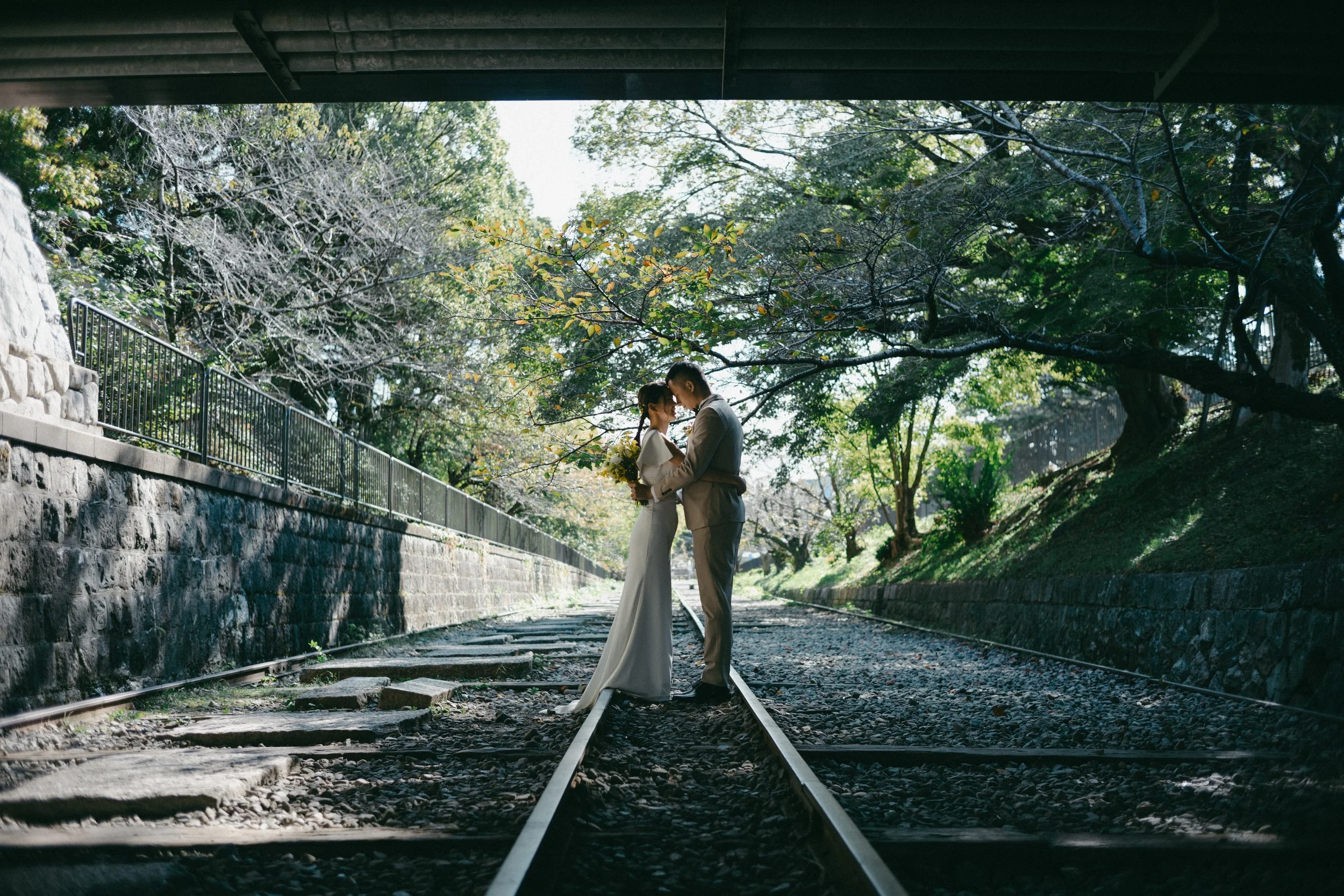 A bride and groom standing close together on a railway track under a bridge, with trees and sunlight in the background.