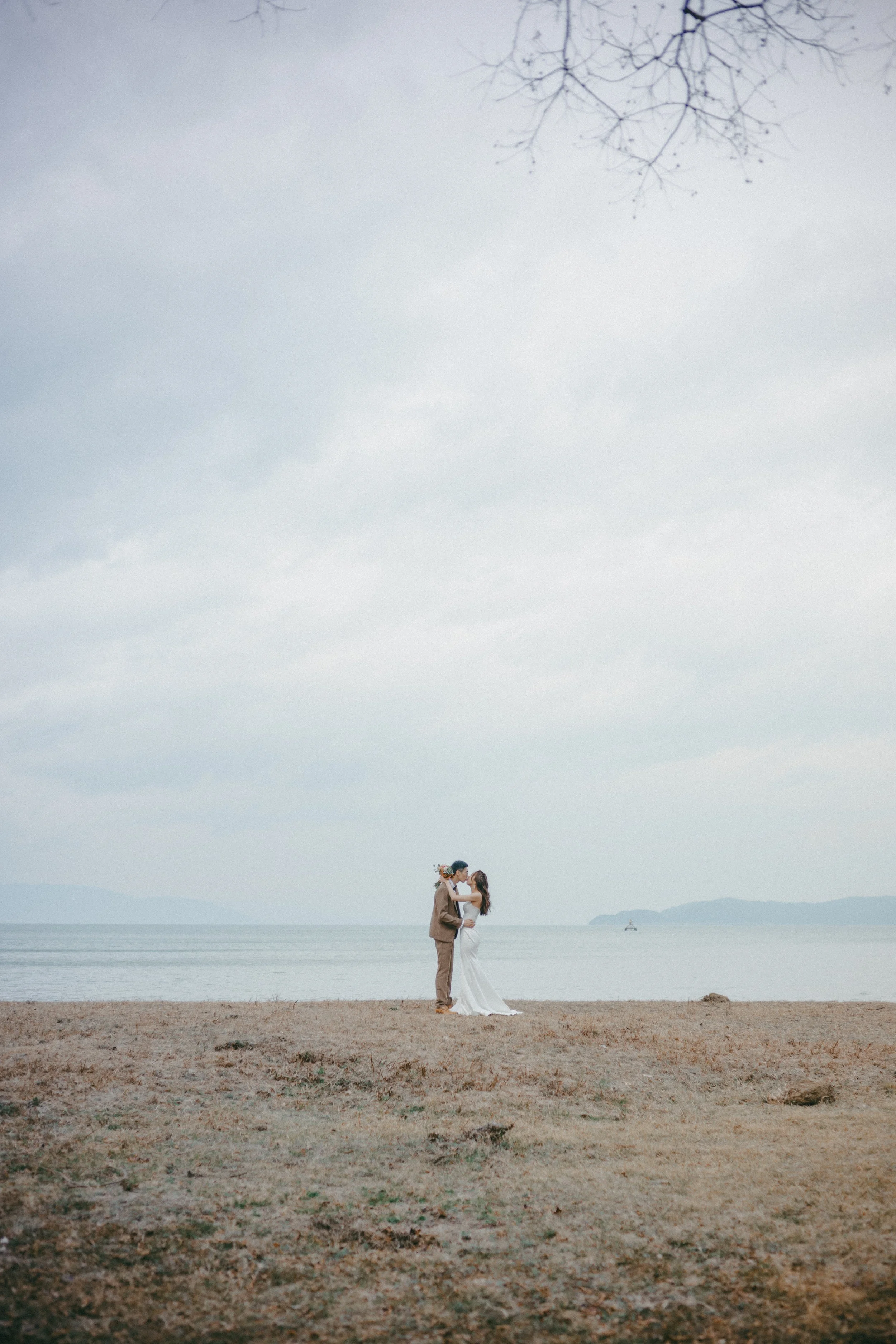 Couple in wedding attire standing on a beach, embracing near the water with overcast sky and distant mountains in the background.