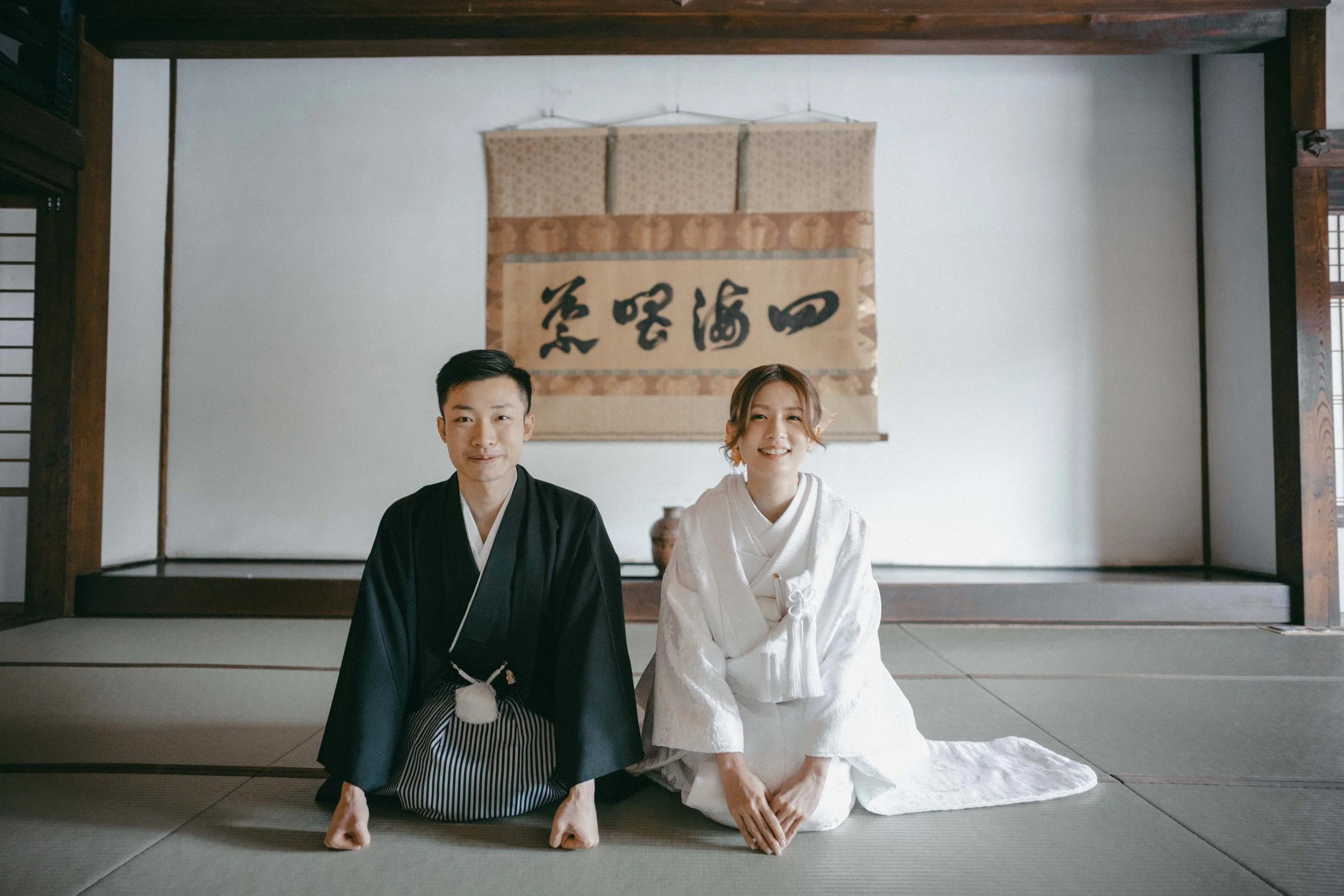 A couple dressed in traditional Japanese wedding attire, kneeling on tatami mats inside a traditional room with a hanging calligraphy scroll in the background.
