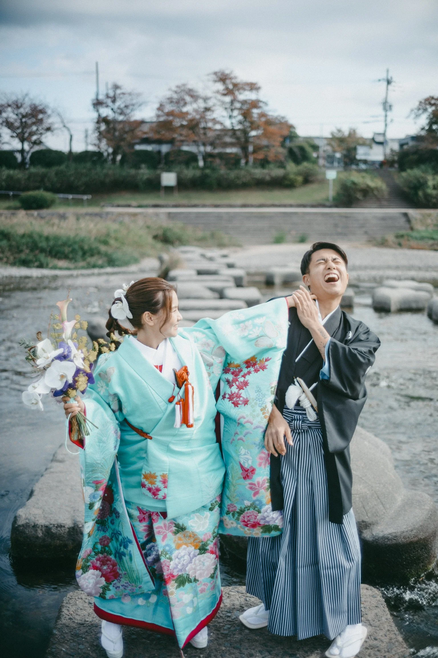 A couple dressed in traditional Japanese wedding attire standing on rocks in a river, with the bride holding a bouquet and the groom expressing pain or discomfort.