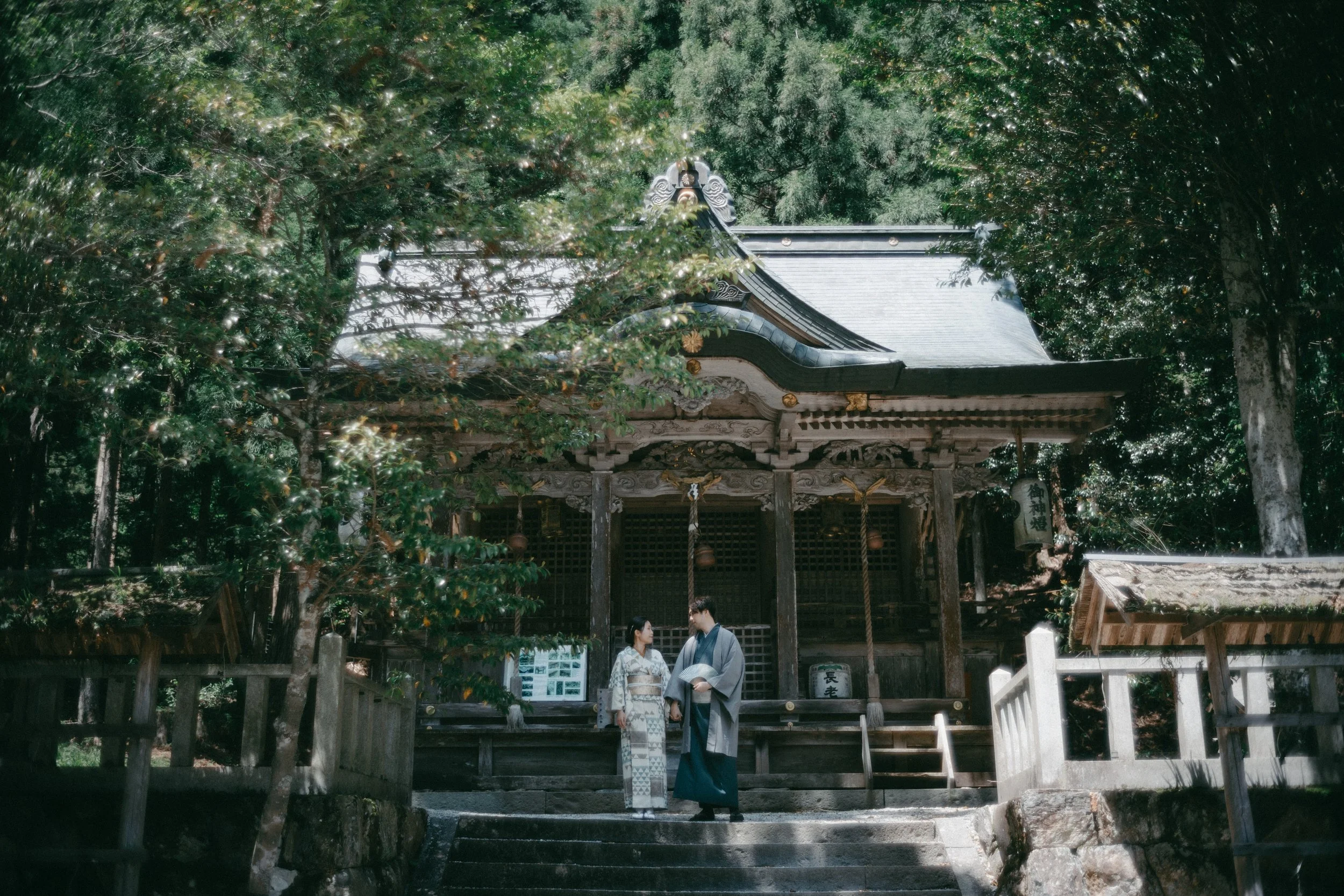 A couple dressed in traditional Japanese clothing standing on temple steps, surrounded by trees and a wooden temple structure.