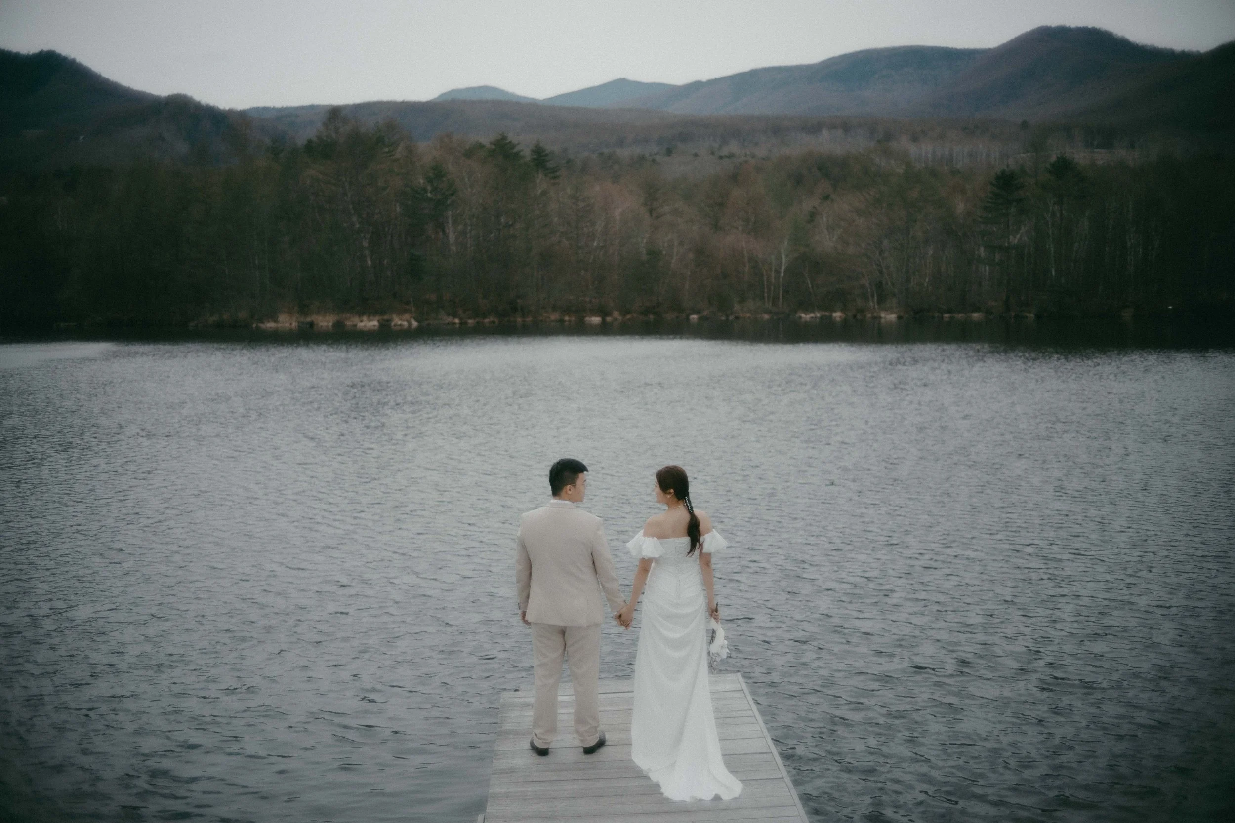 A man and woman in wedding attire standing hand in hand on a dock by a lake, facing away, with mountains and trees in the background.
